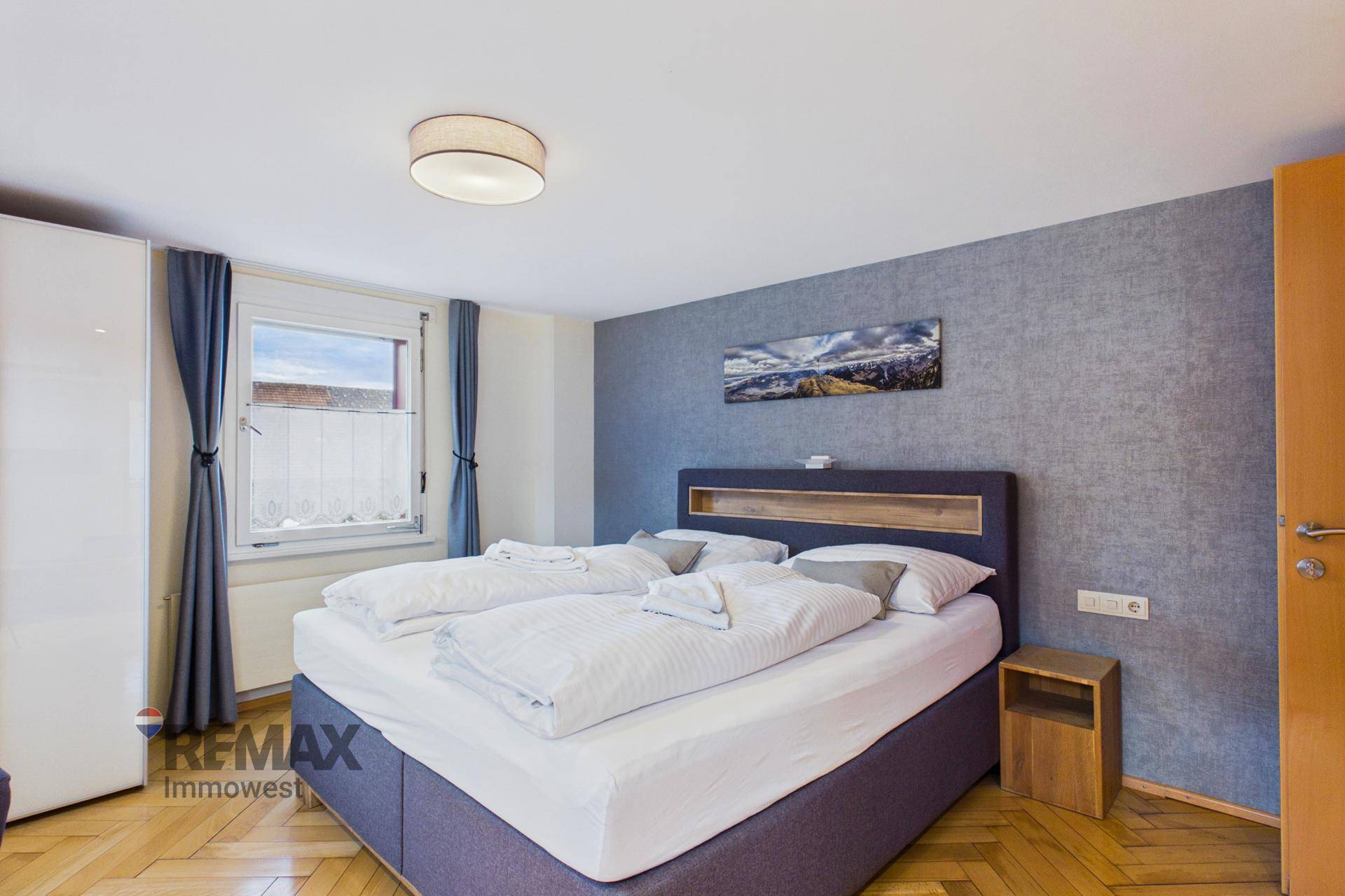 Helles Schlafzimmer mit Doppelbett, Holzboden und einem Fenster mit Vorhängen.