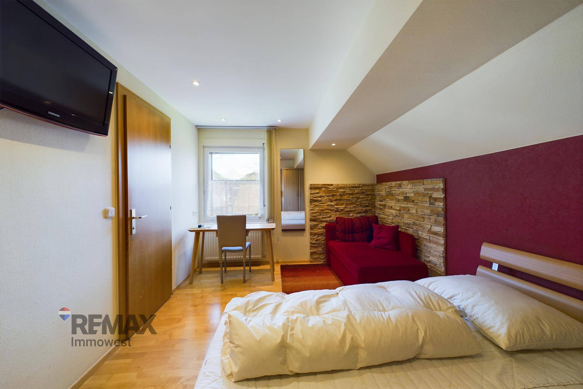 Schlafzimmer mit Holzboden, roter Akzentwand, Schreibtisch und Dachschräge.