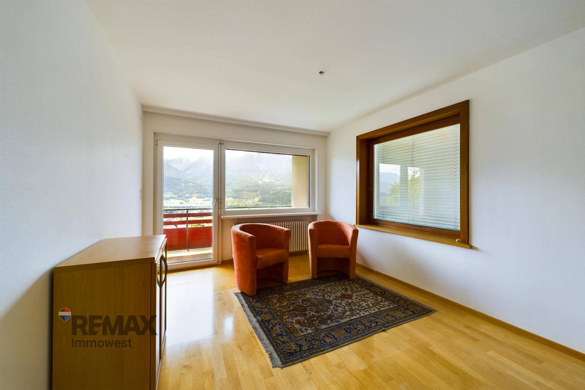 Heller Raum mit Holzboden, zwei orangefarbenen Sesseln und großem Fenster mit Bergblick.