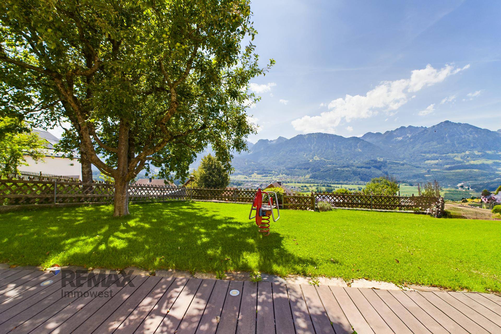 Großer Gartenbereich mit Rasenfläche, Baum und Kinderspielgerät, umgeben von Bergen.