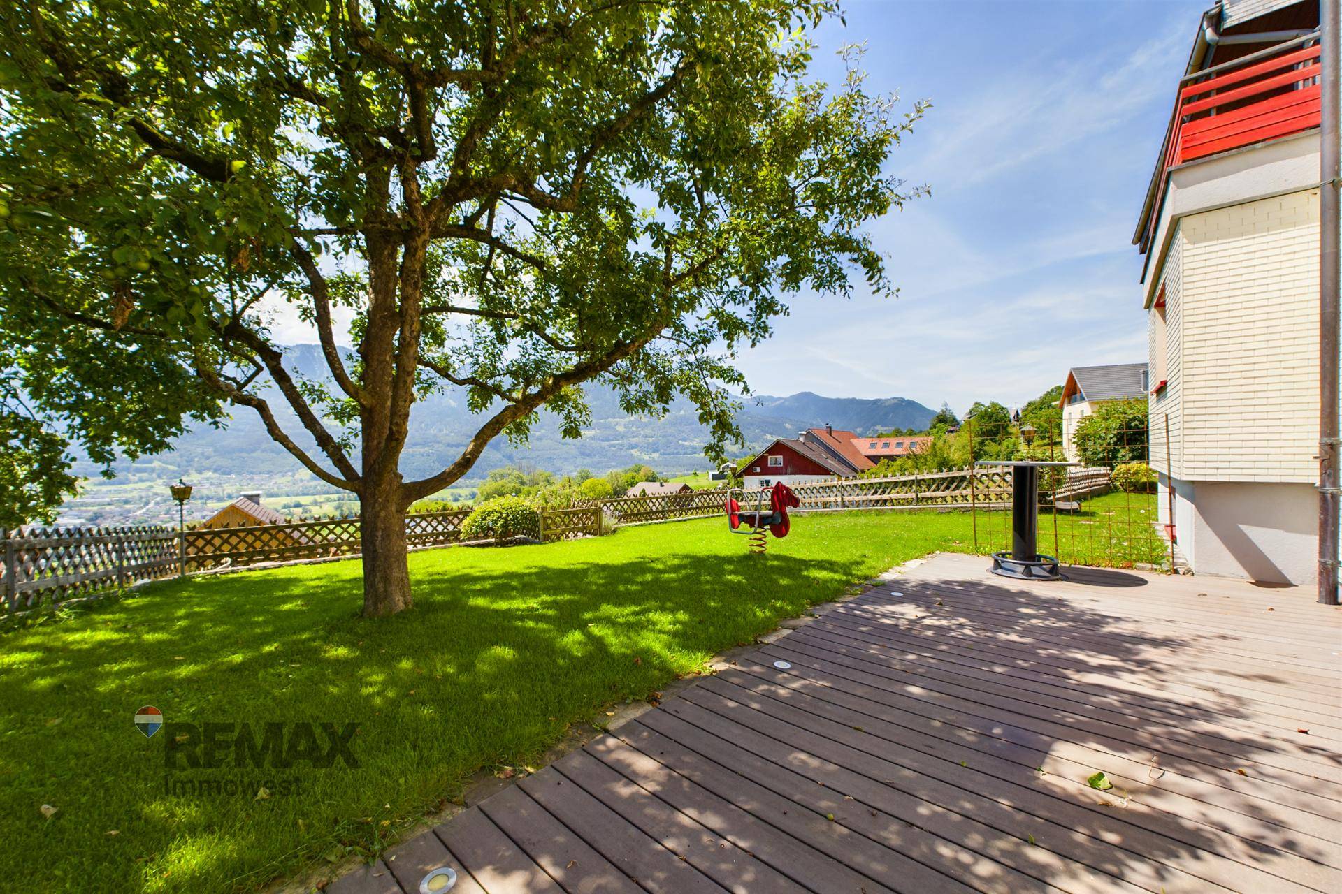 Garten mit Holzterrasse, großem Baum und Kinderspielgerät, umgeben von Natur.