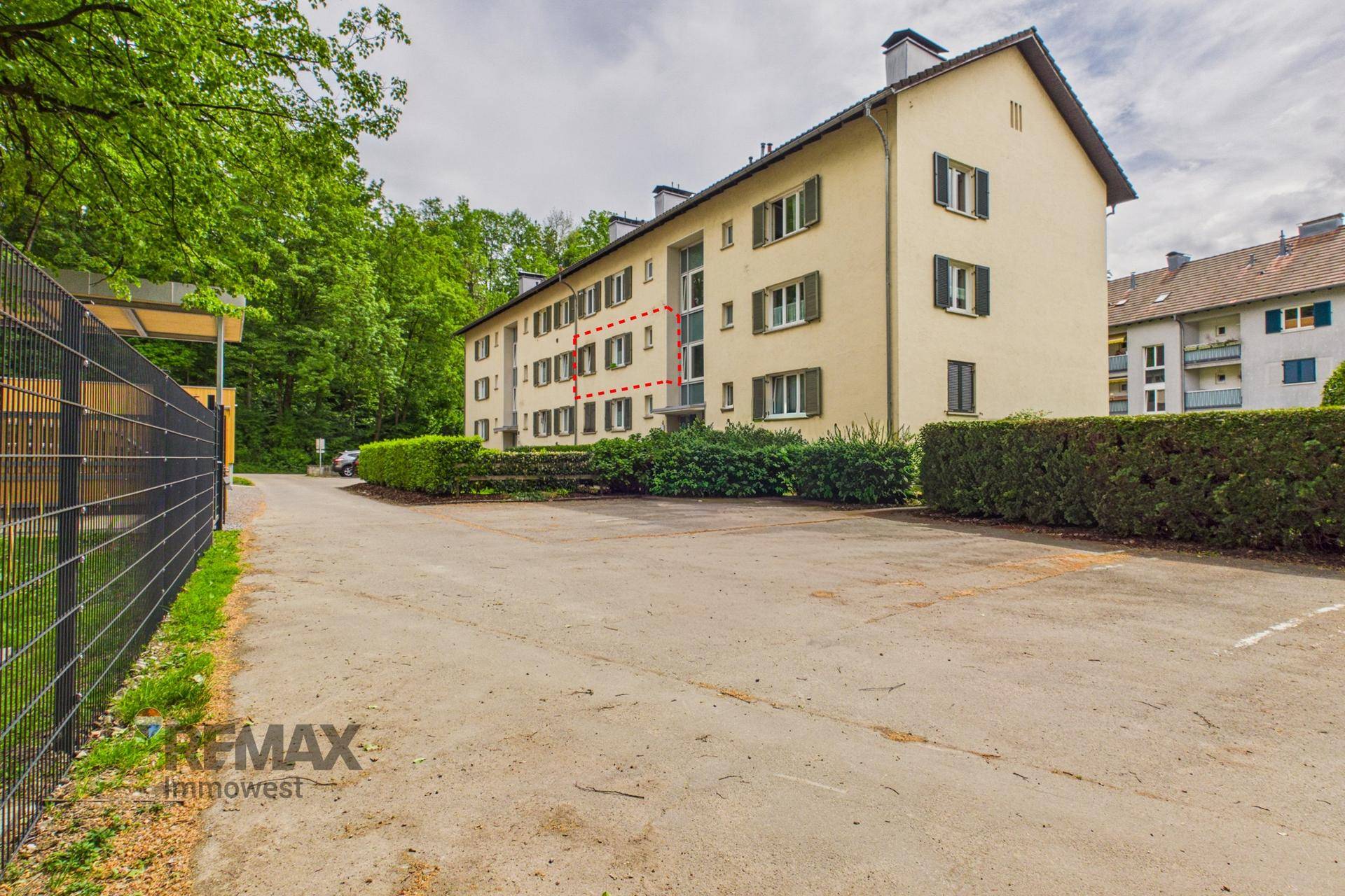 Seitenansicht des Mehrfamilienhauses mit Parkplatz und angrenzendem Waldrand.