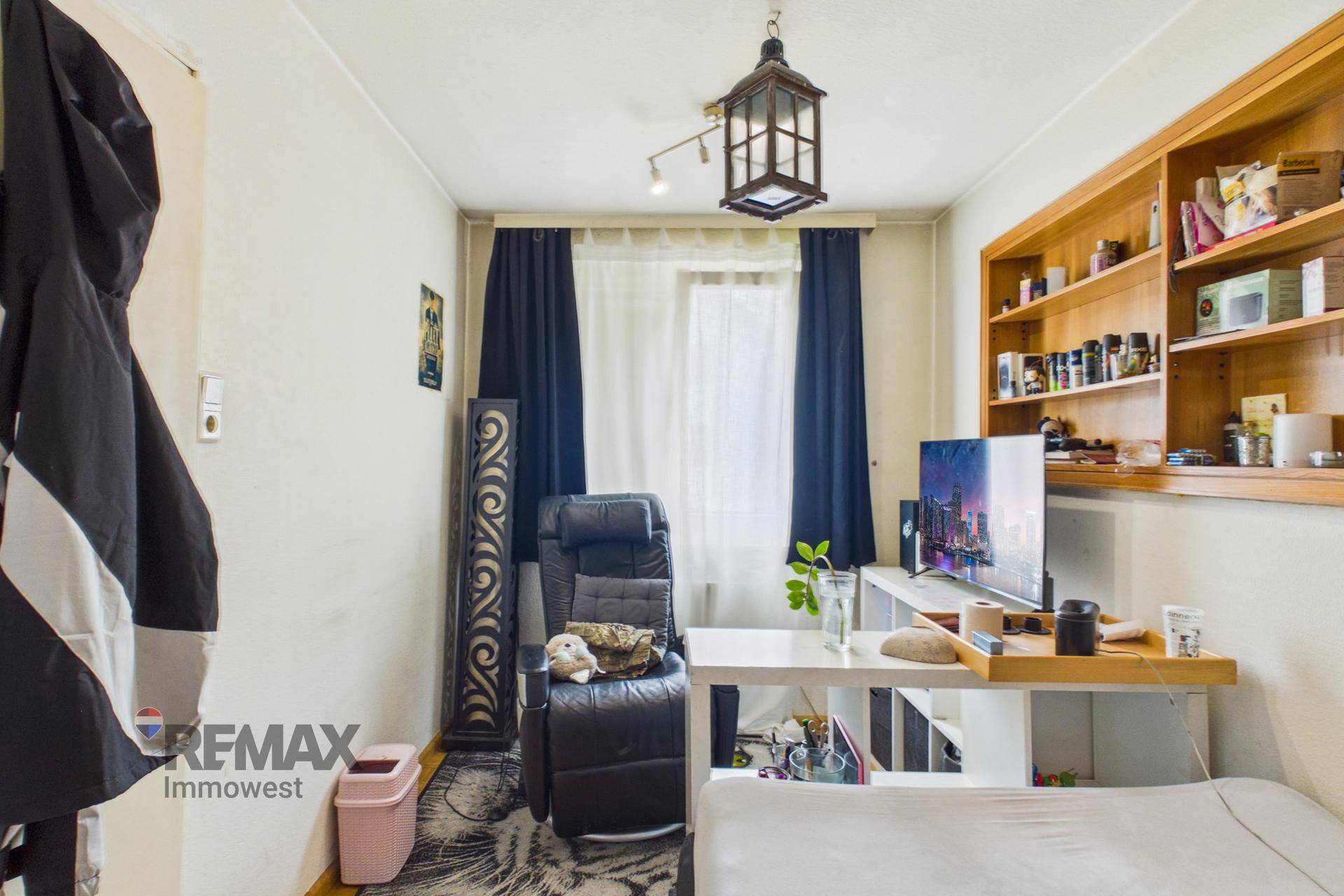 Kleines Arbeitszimmer mit Schreibtisch, Regal und dunklen Vorhängen.