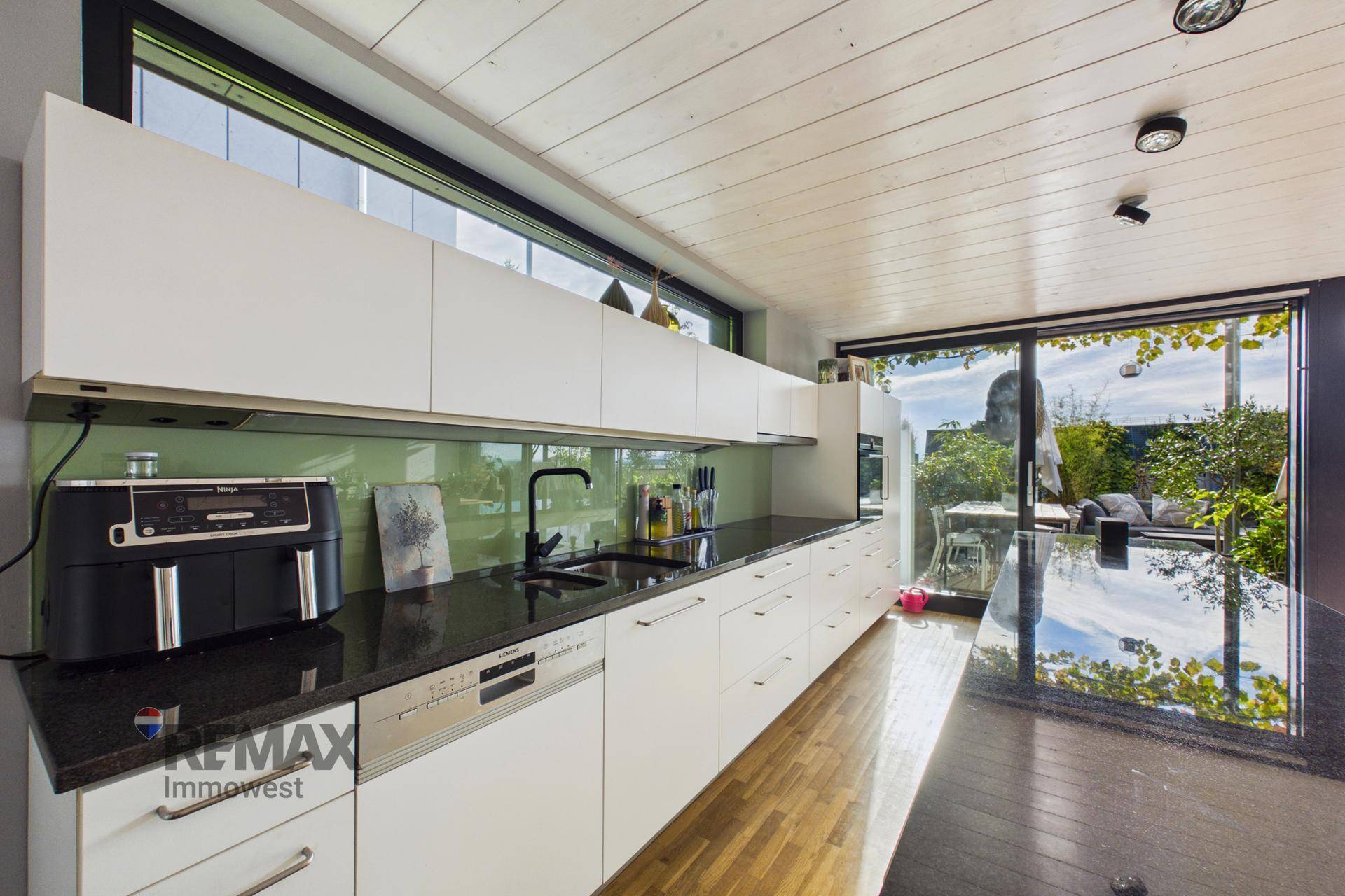 Moderne Küche mit weißen Schränken, dunkler Arbeitsplatte und großem Fenster mit Blick ins Grüne.