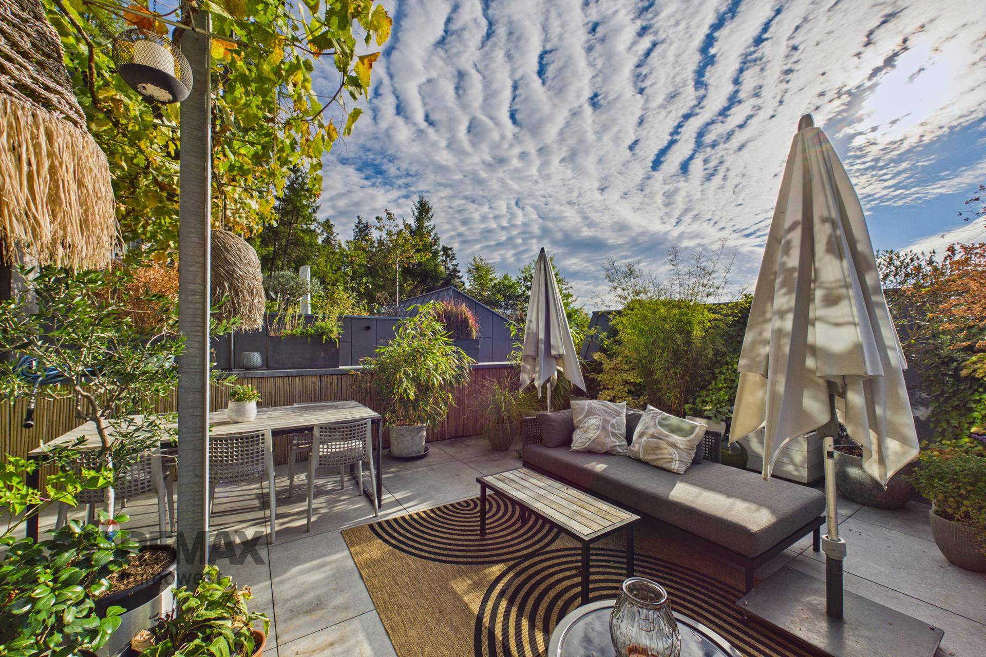 Sonniger Terrassenbereich mit Lounge-Möbeln, Esstisch und üppiger Bepflanzung unter blauem Himmel.