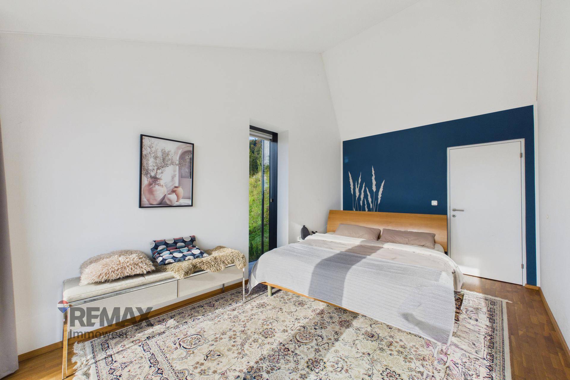 Schlafzimmer mit Holzboden, blauer Akzentwand und Fenster mit Blick ins Grüne.