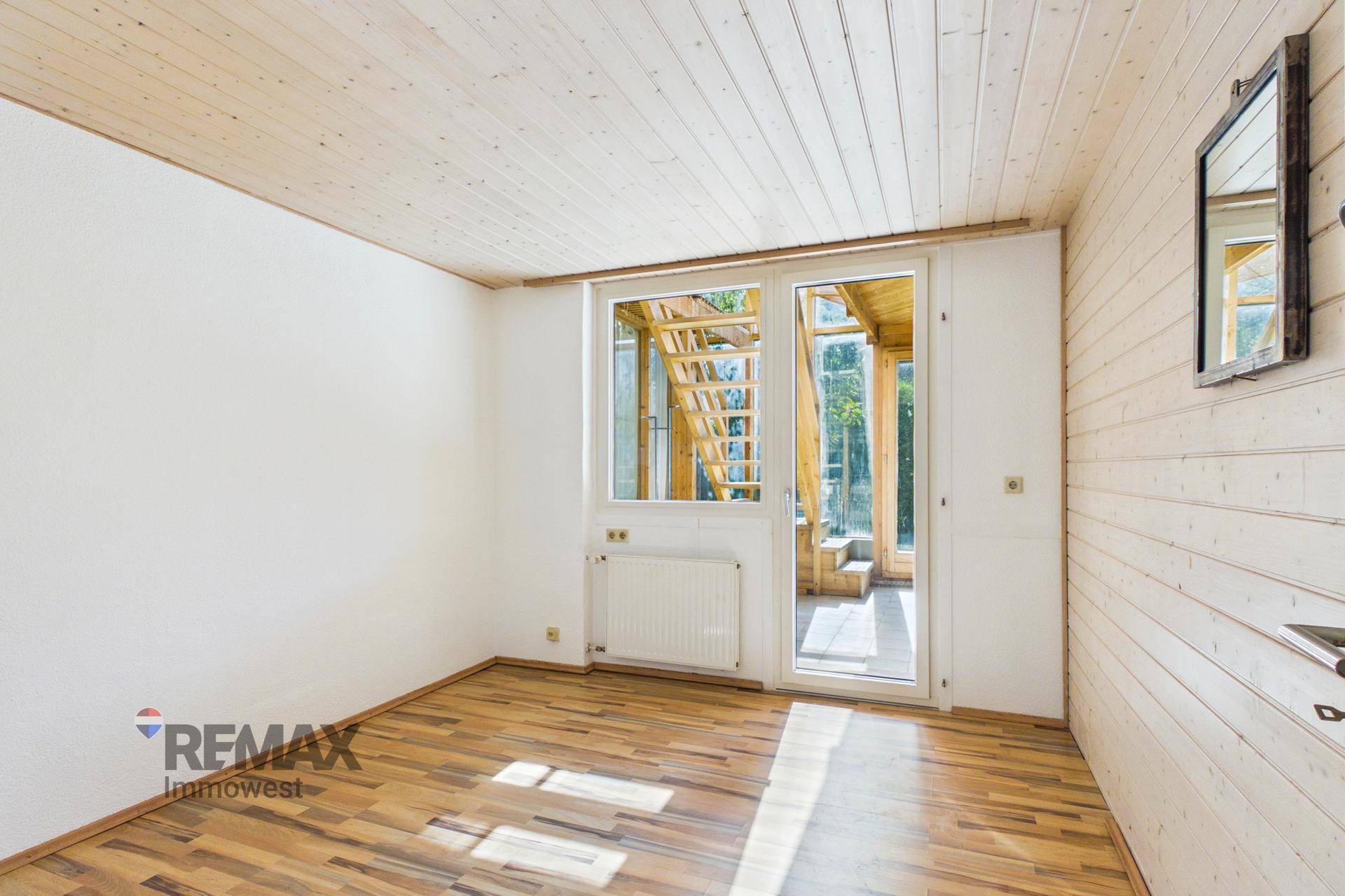 Leerer Raum mit Holzverkleidung, Holzboden und Zugang zu einem verglasten Anbau, der viel Licht bietet.