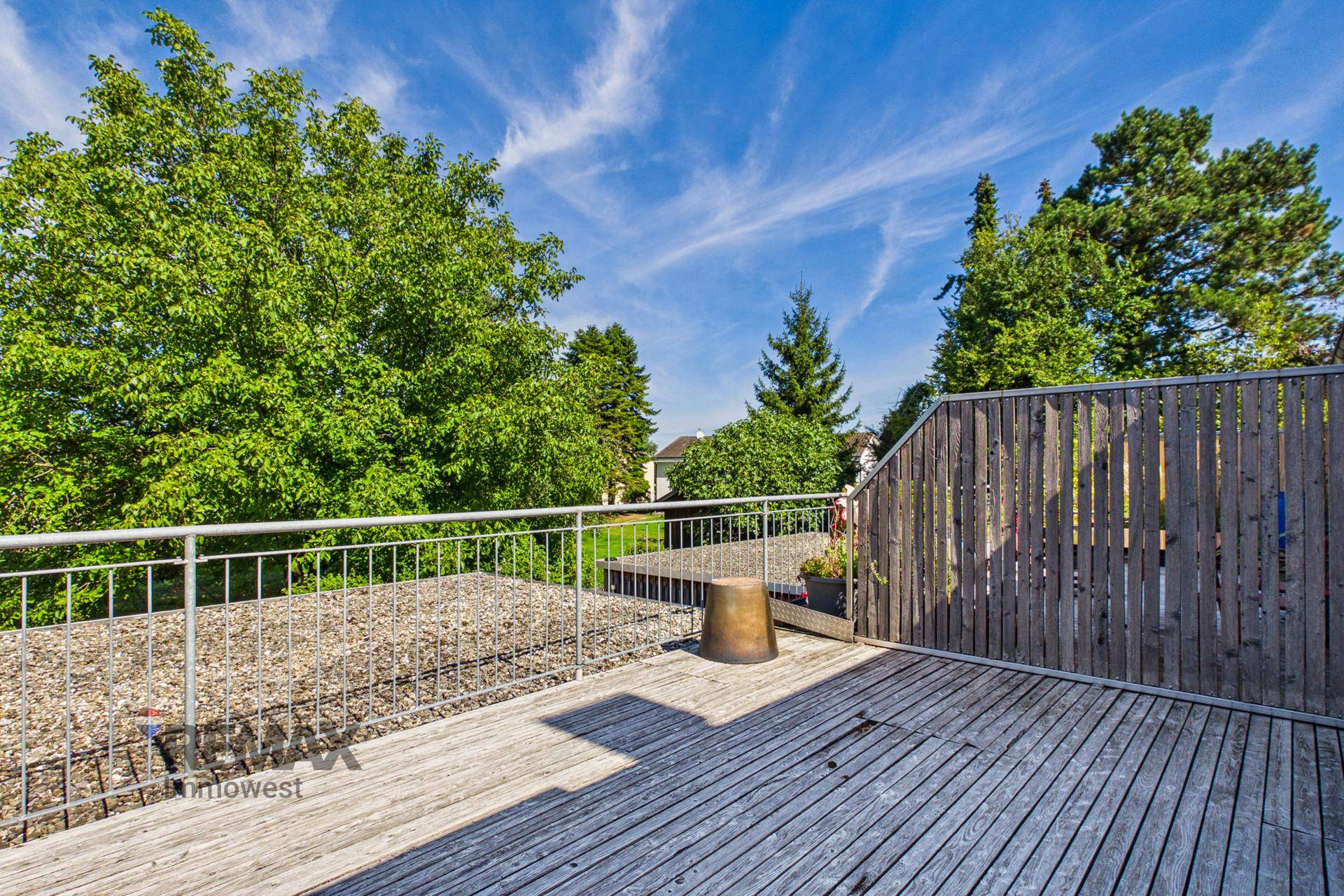 Große Holzterrasse mit Metallgeländer und Blick auf Bäume unter blauem Himmel, ideal für Entspannung.