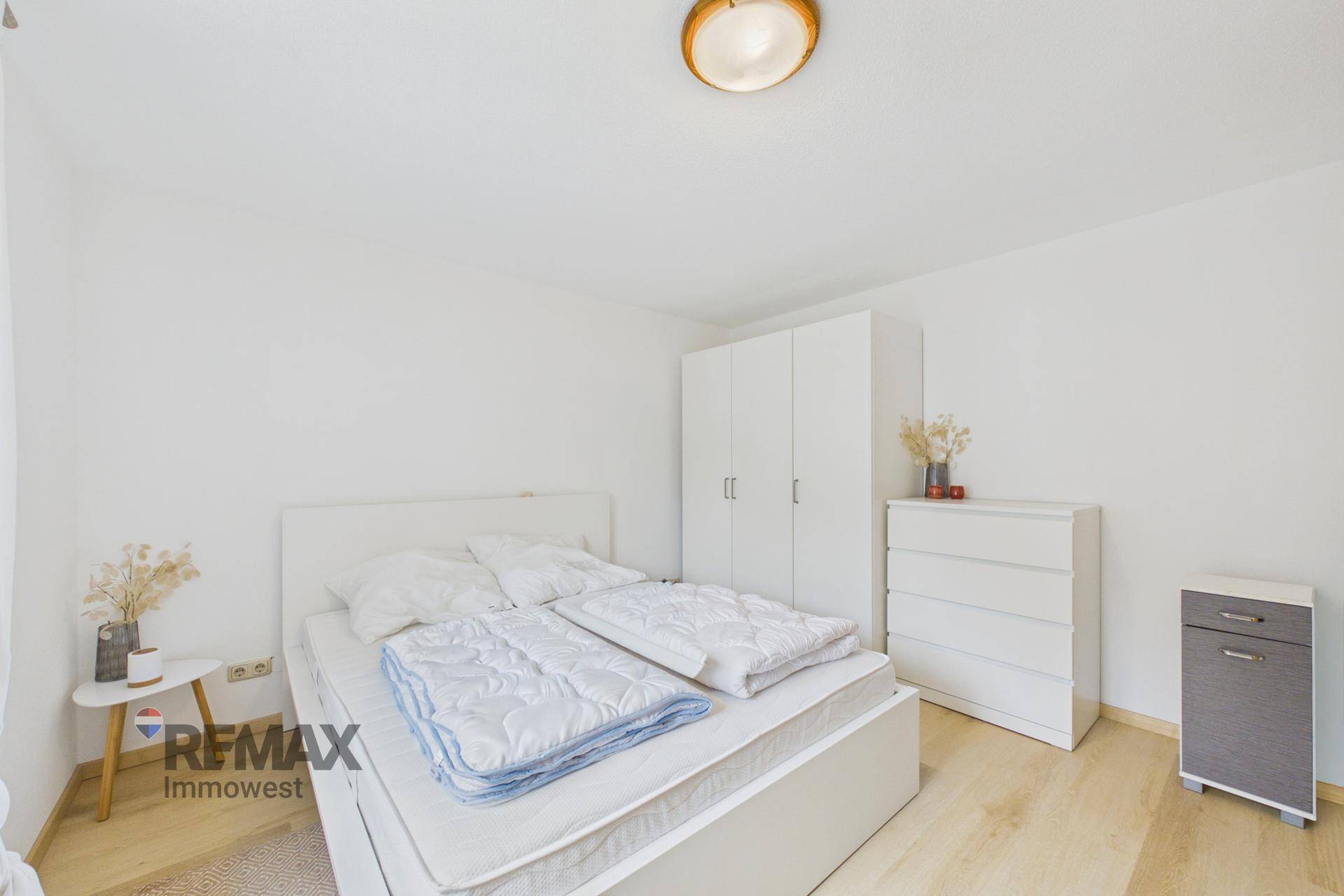Schlafzimmer mit weißem Bett, Kleiderschrank und Kommode auf hellem Holzfußboden.