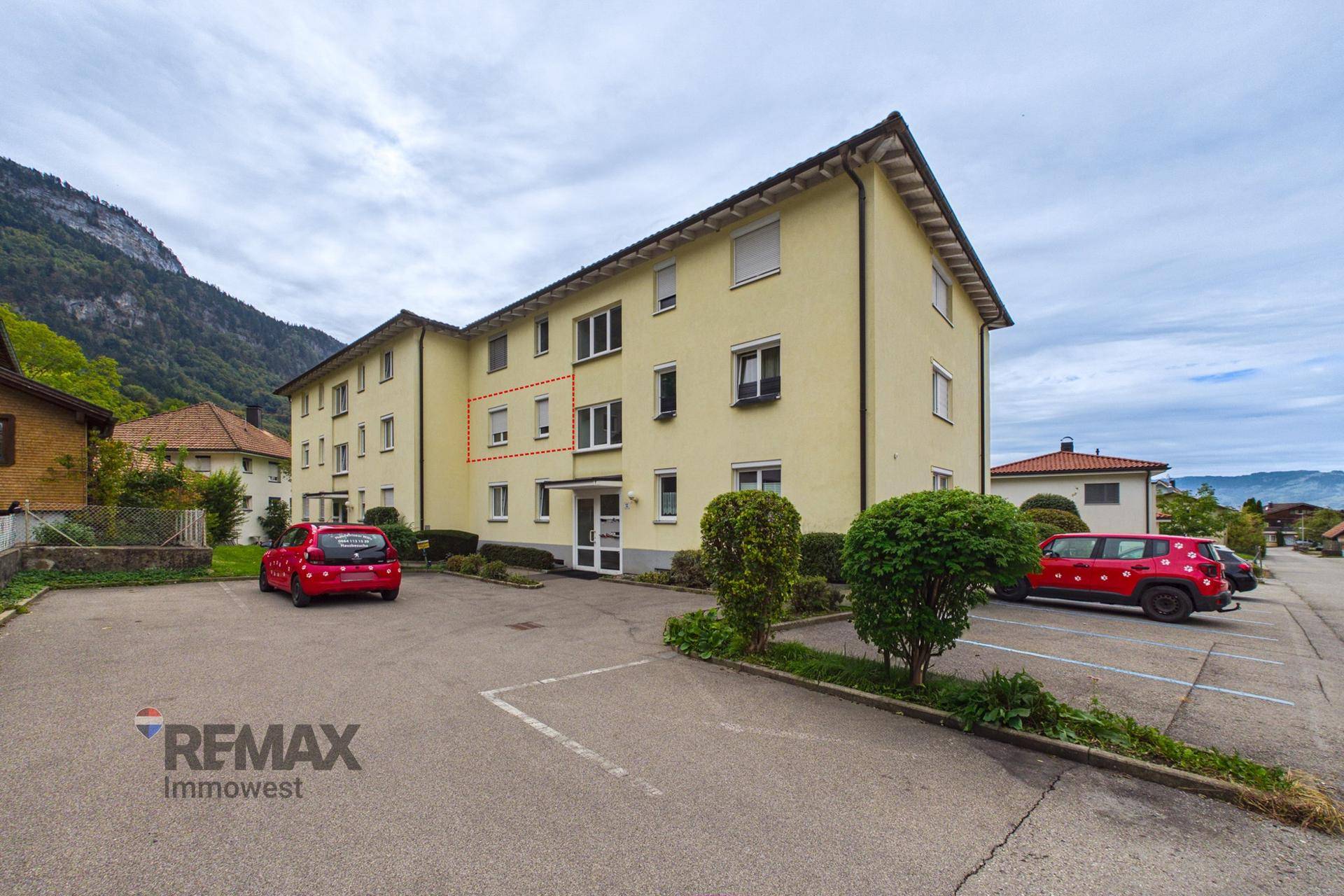 Mehrfamilienhaus mit gelber Fassade und Parkplätzen vor einer Bergkulisse.