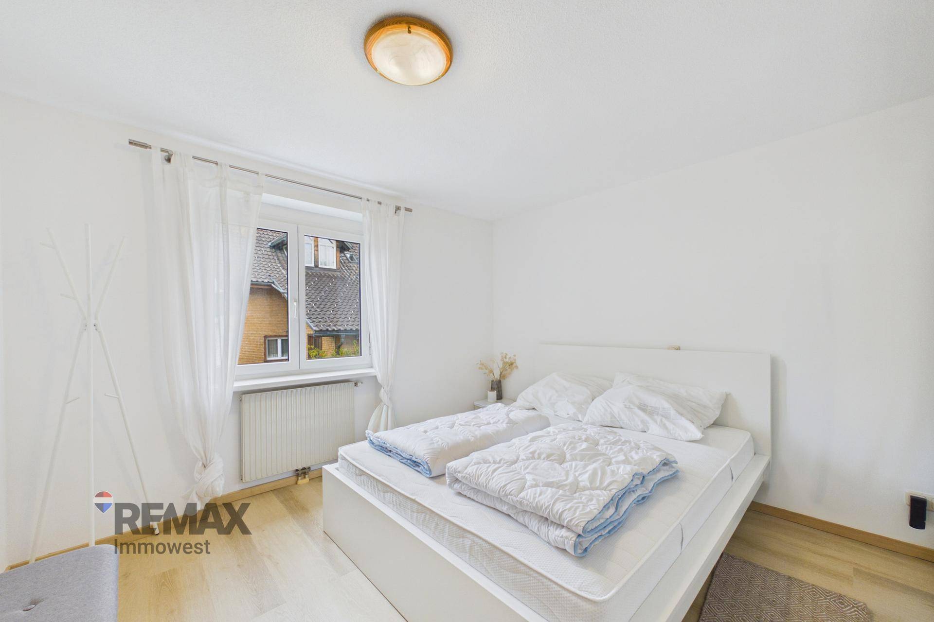 Helles Schlafzimmer mit weißem Bett, Fenster und Heizkörper, ausgestattet mit Vorhängen.