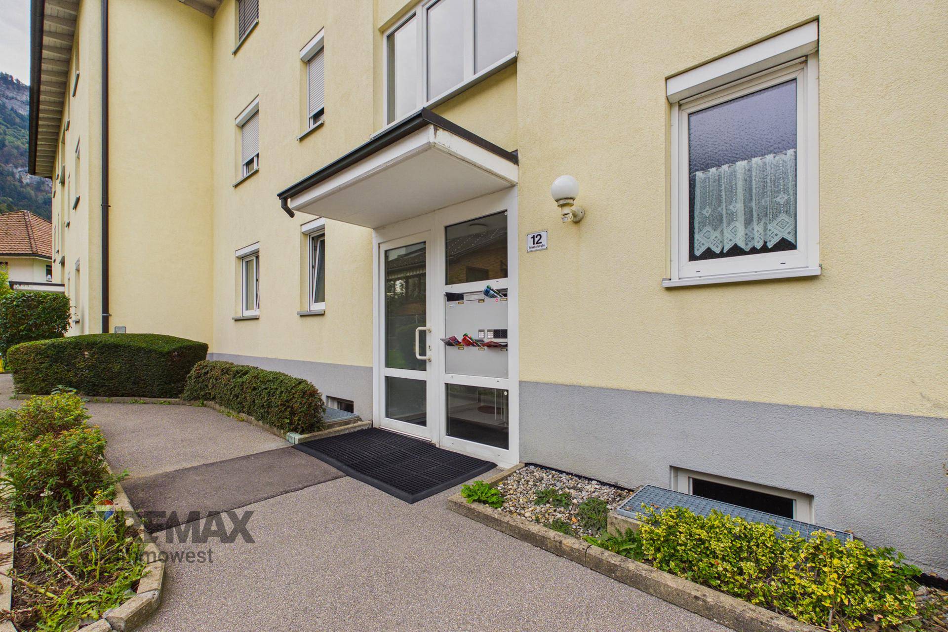 Eingangsbereich eines Mehrfamilienhauses mit Glasportal und Vordach, umgeben von gepflegten Büschen.