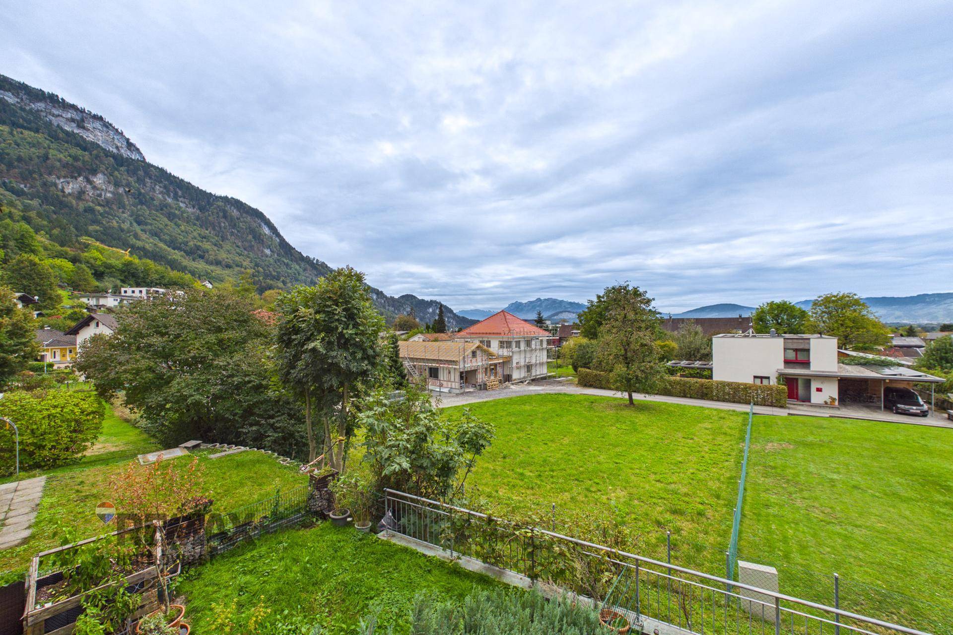 Weitläufiger Blick über grüne Wiesen und Häuser mit Bergen im Hintergrund unter bewölktem Himmel.