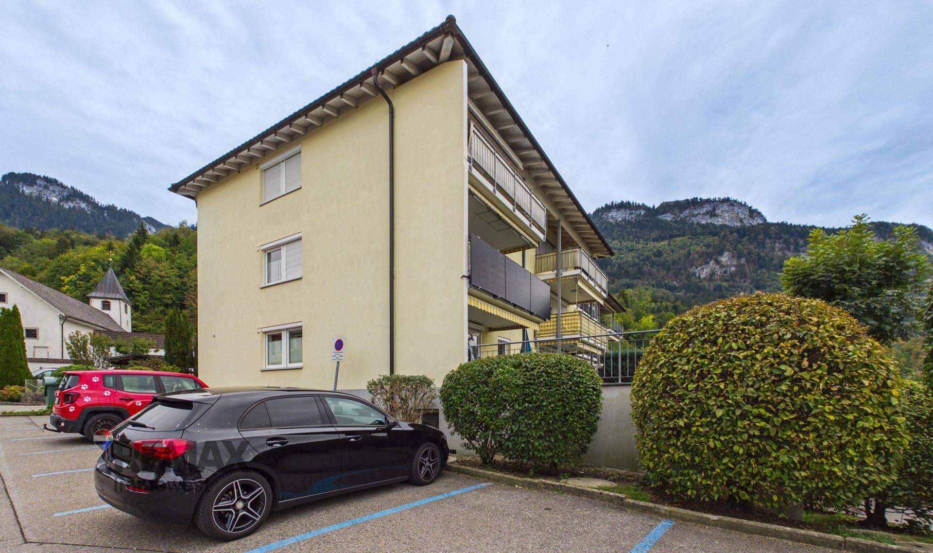 Seitenansicht des Mehrfamilienhauses mit Parkplätzen und Blick auf die Berge.
