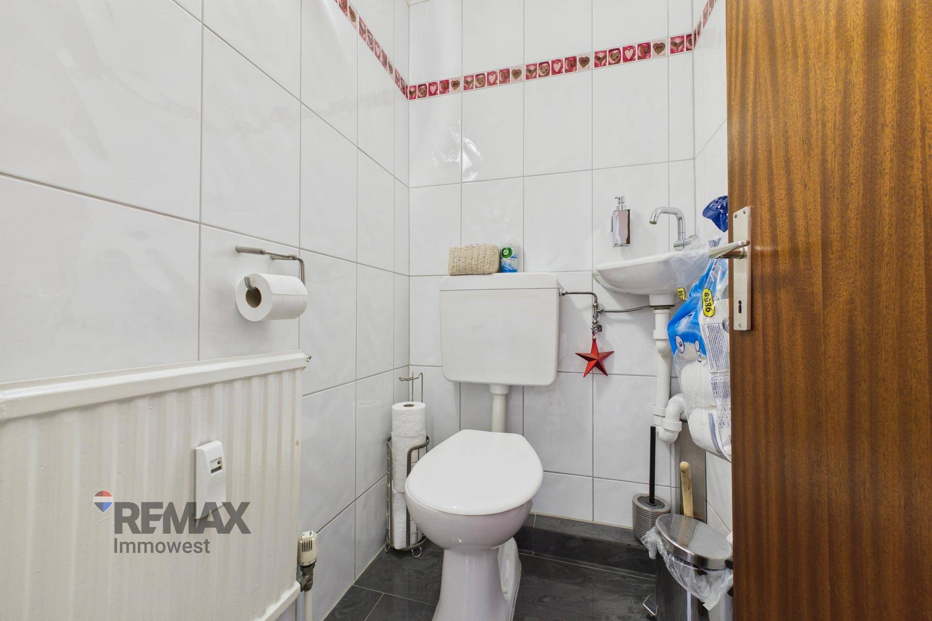 Kleines Gäste-WC mit Toilette, Handwaschbecken und weißen Fliesen mit roter Bordüre.
