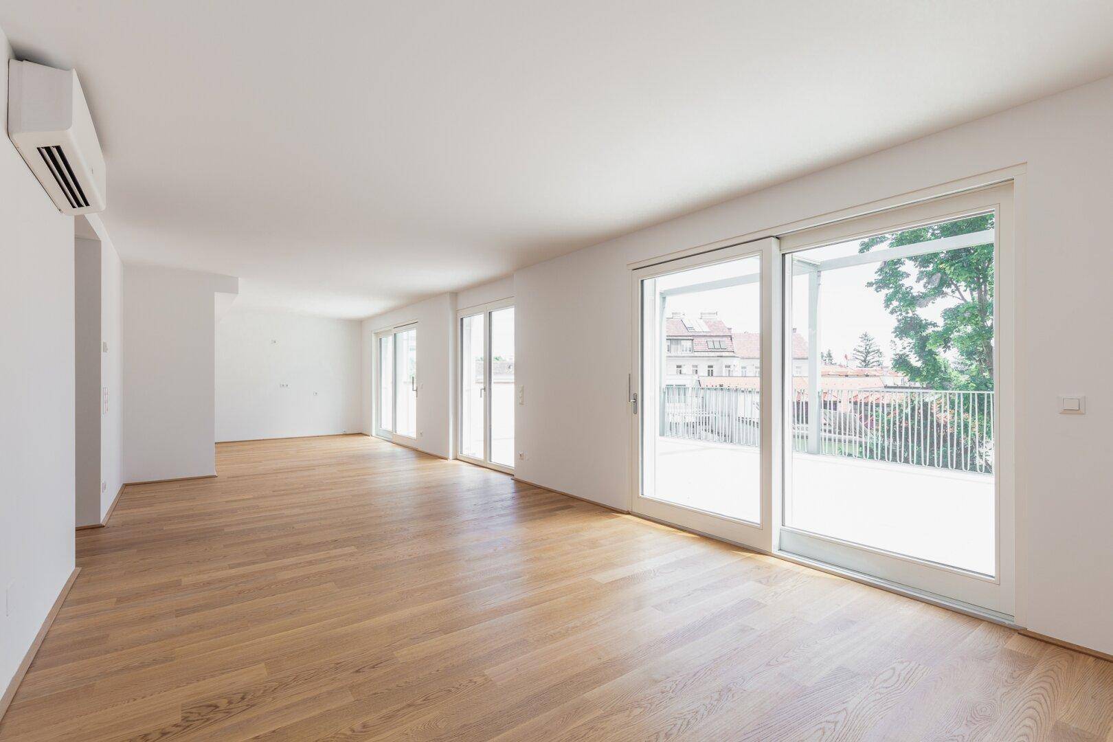 Heller Wohnbereich mit Holzboden, Klimaanlage und großen Fenstern mit Blick ins Grüne.