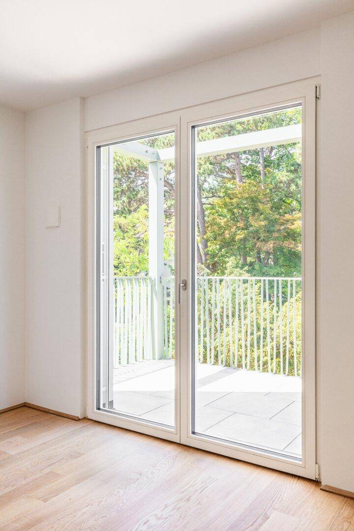 Helle Fenstertüren führen auf einen Balkon mit Blick auf Bäume und Grünflächen.