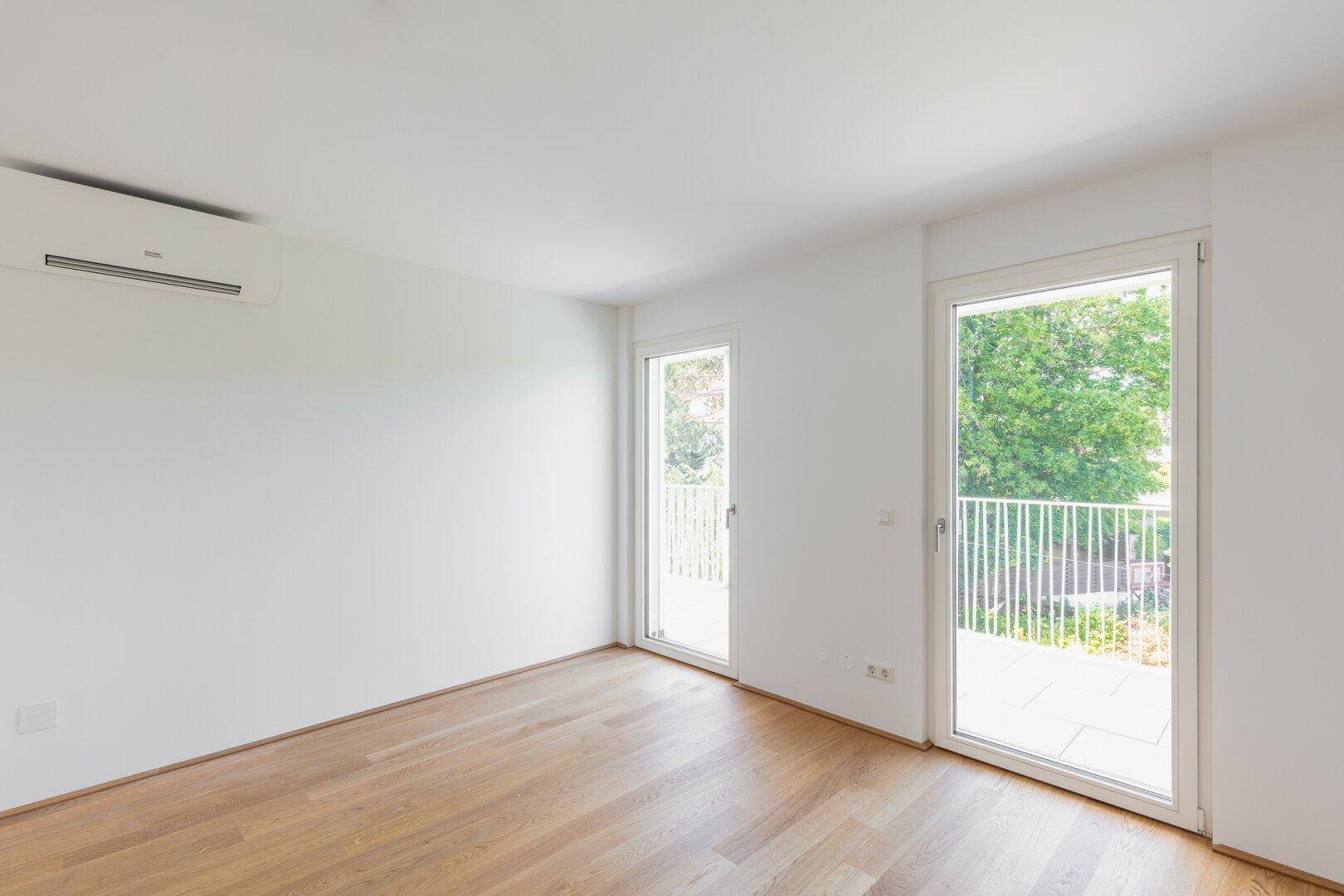 Helles Zimmer mit Holzboden, Klimaanlage und zwei Fenstertüren zum Balkon.