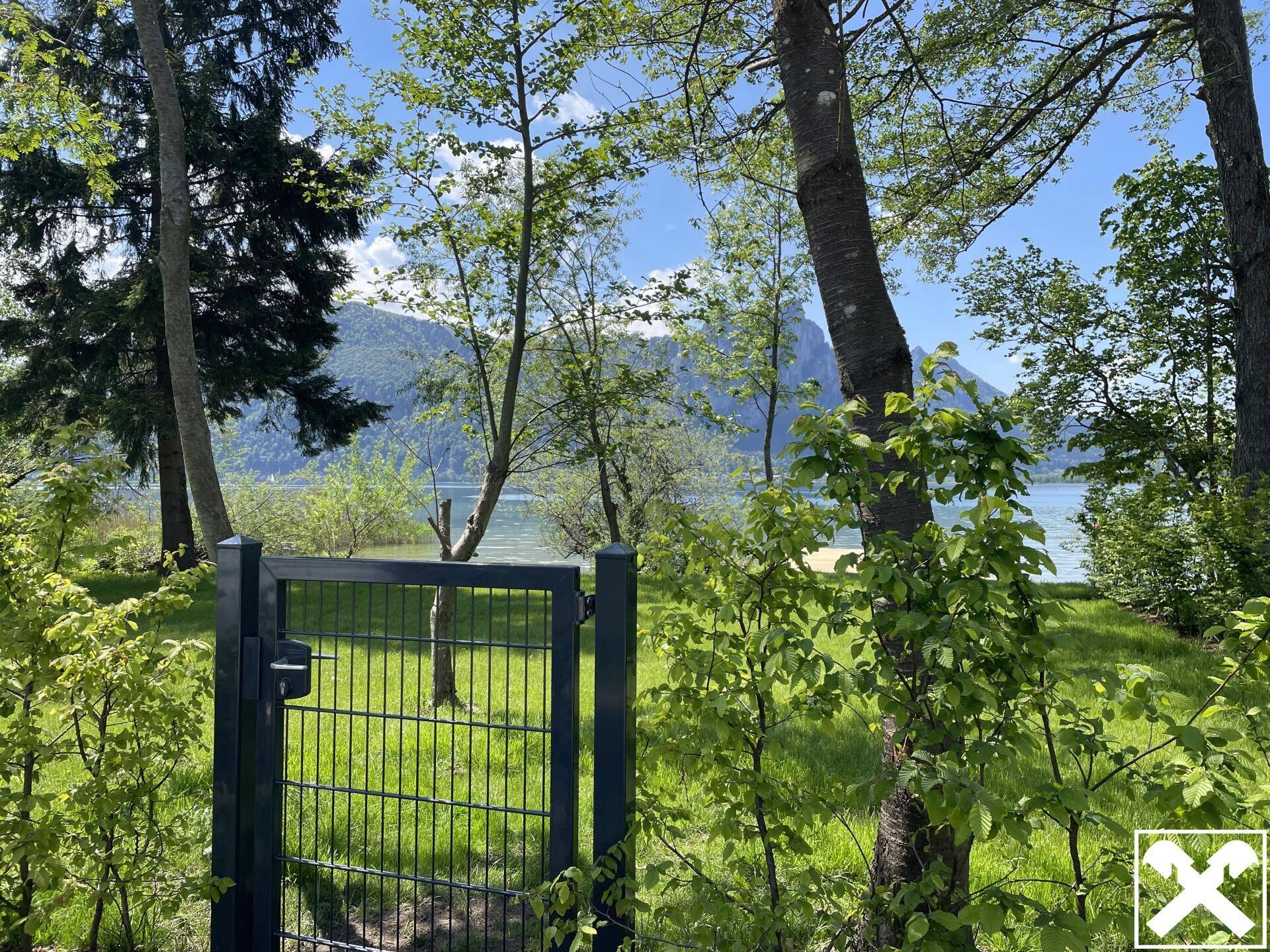Gartentor umgeben von Bäumen, das den Zugang zum See mit Bergblick an einem hellen Tag ermöglicht.