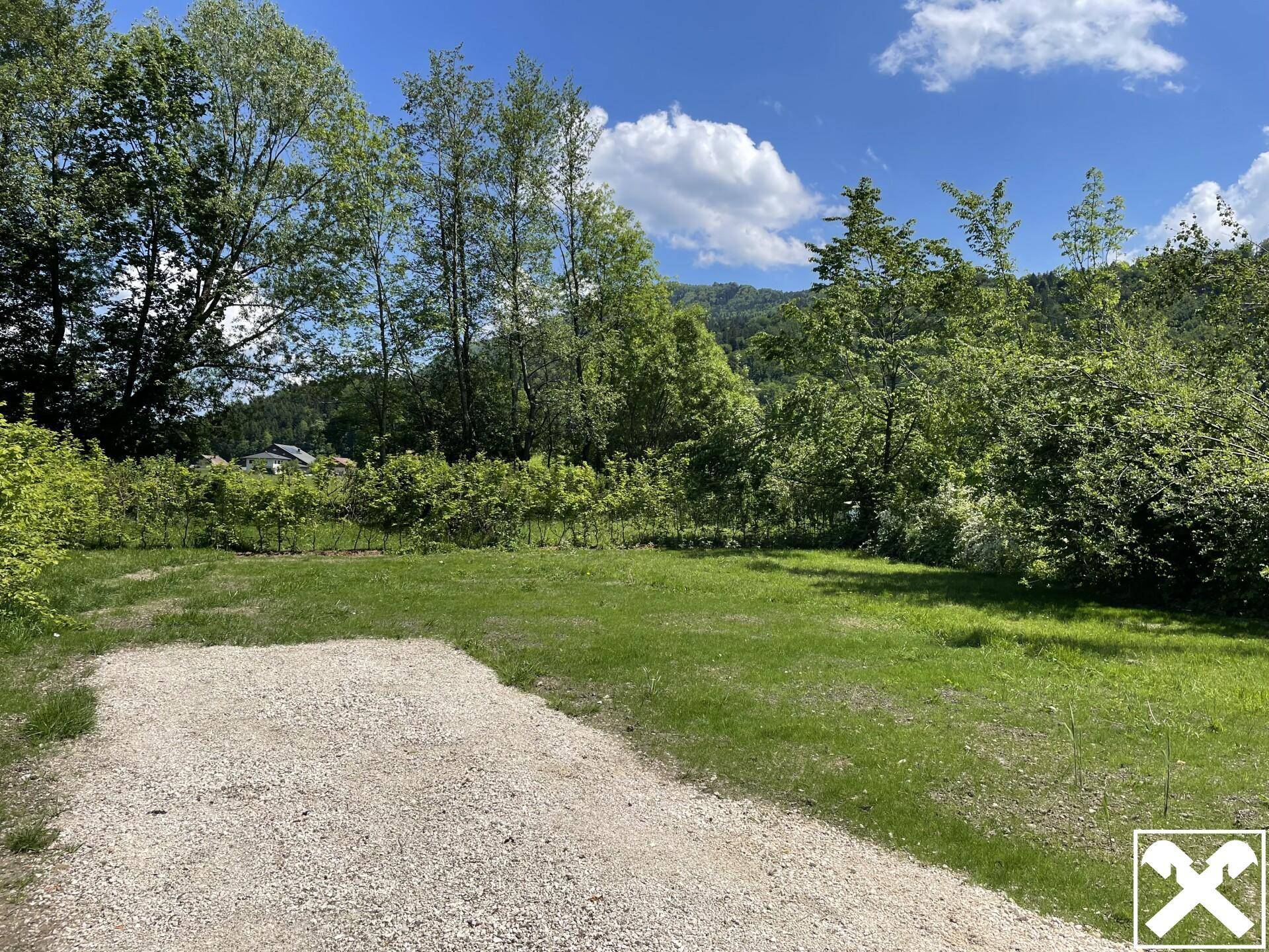 Offene Grünfläche mit Kiesweg, umgeben von Bäumen und Blick auf die Hügellandschaft.