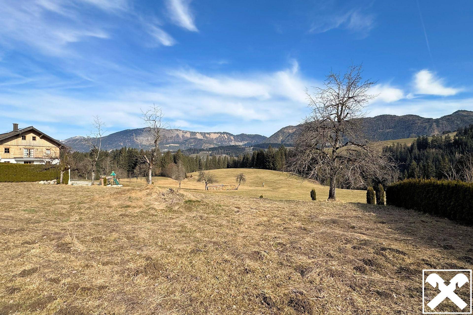 Panoramablick über ein weitläufiges Feld mit einem traditionellen Haus und der beeindruckenden Bergkulisse.