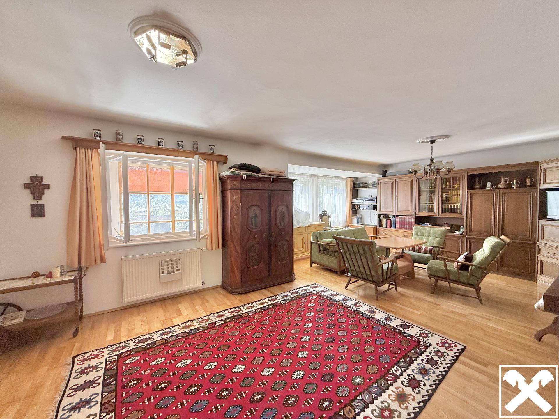 Gemütliches Wohnzimmer mit traditionellen Holzmöbeln, einem großen Teppich und viel Tageslicht.