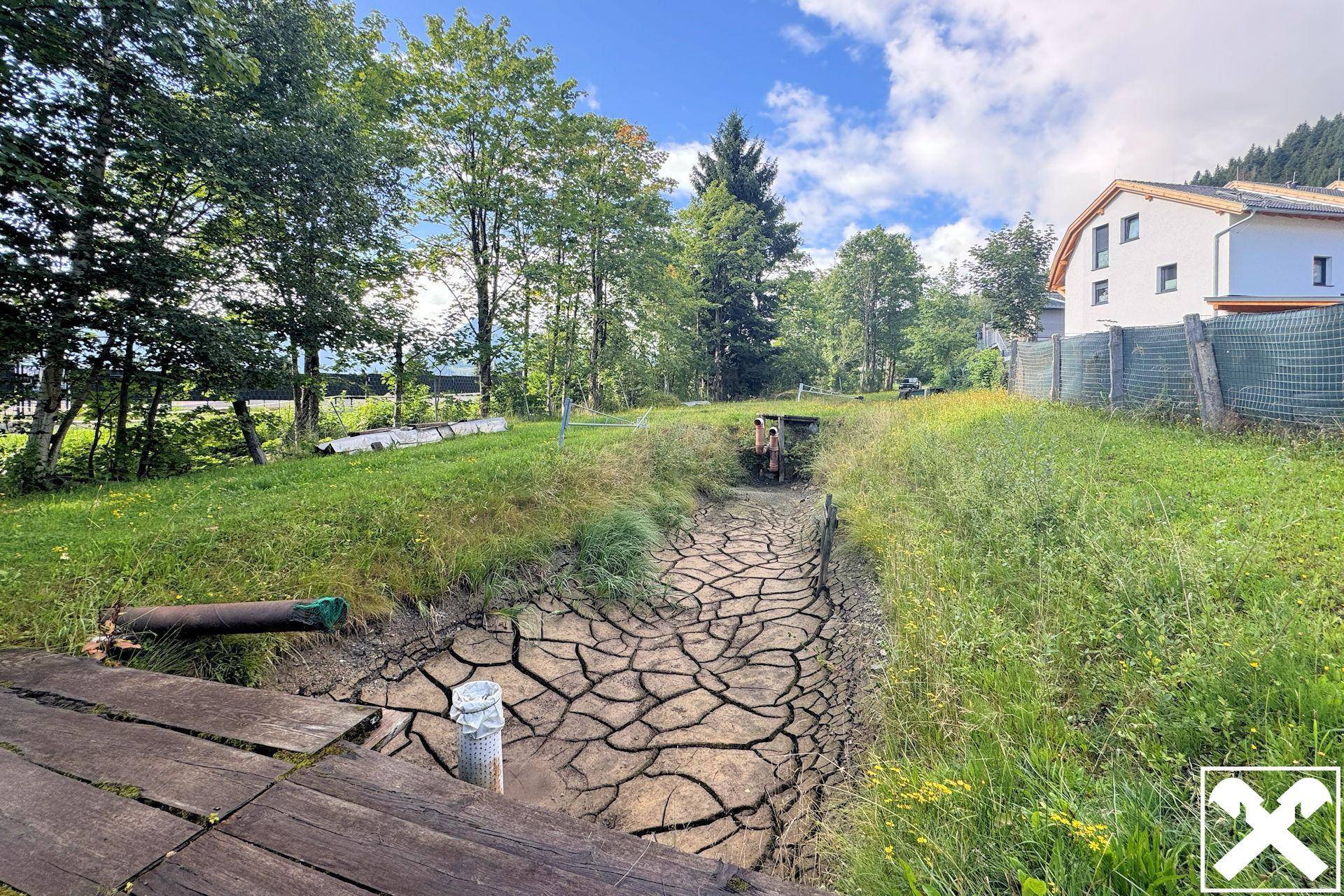 Grünes Grundstück mit einem ausgetrockneten Bachbett und Bäumen im Hintergrund.