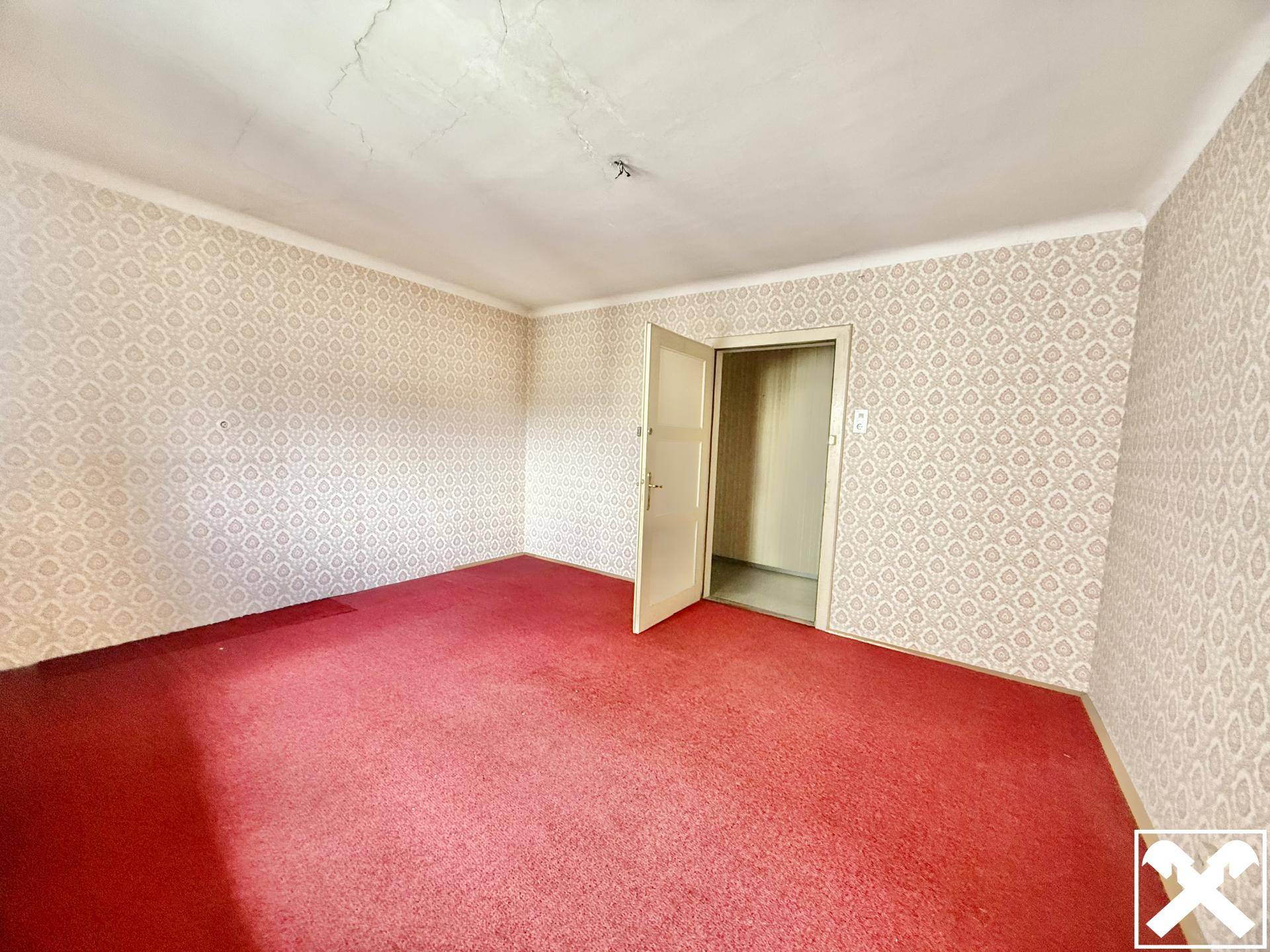 Leeres Zimmer mit rotem Teppichboden und gemusterter Tapete, bereit für eine Renovierung.