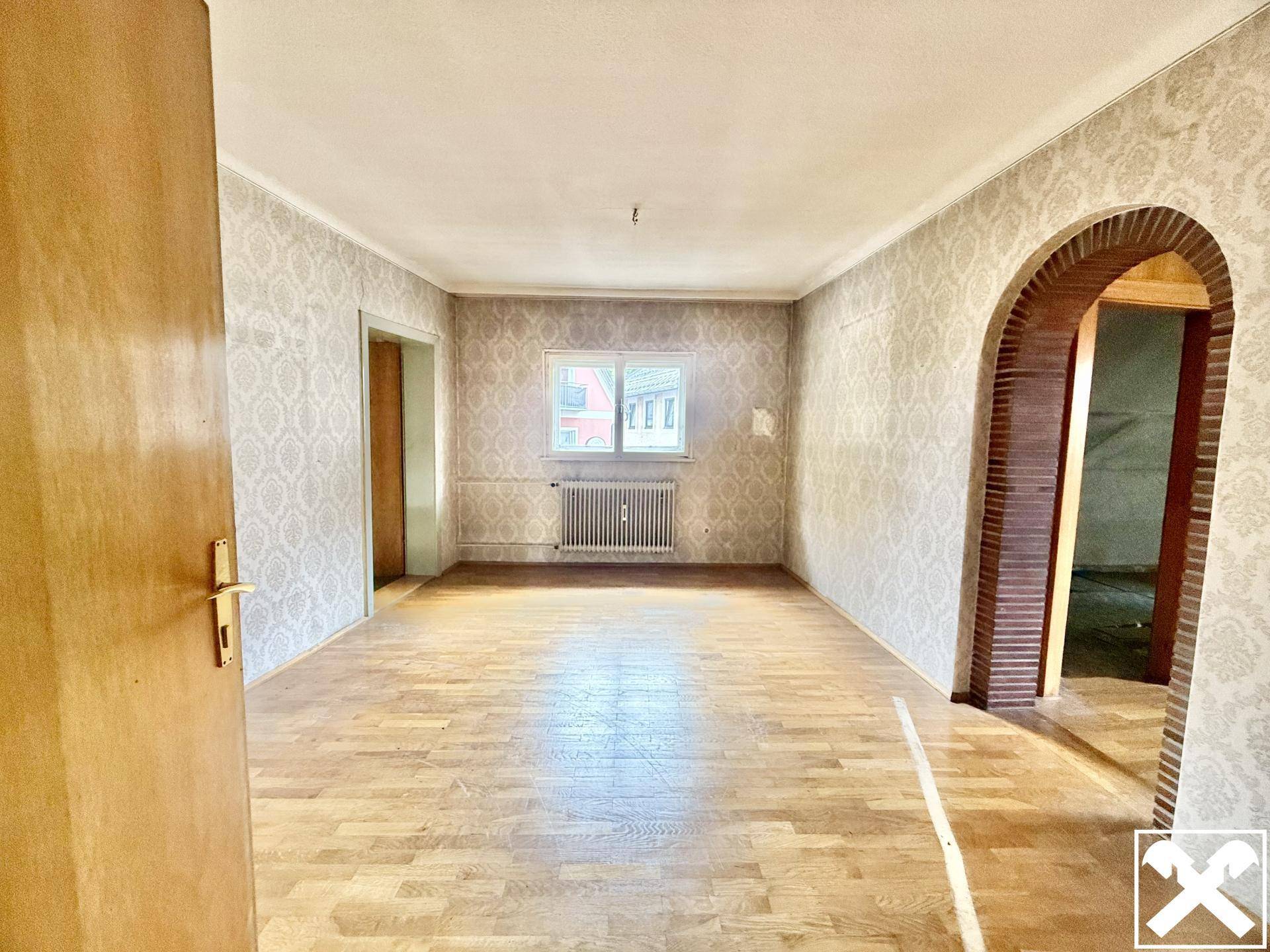 Geräumiges Zimmer mit Parkettboden, gemusterter Tapete und einem Fenster mit Heizkörper.