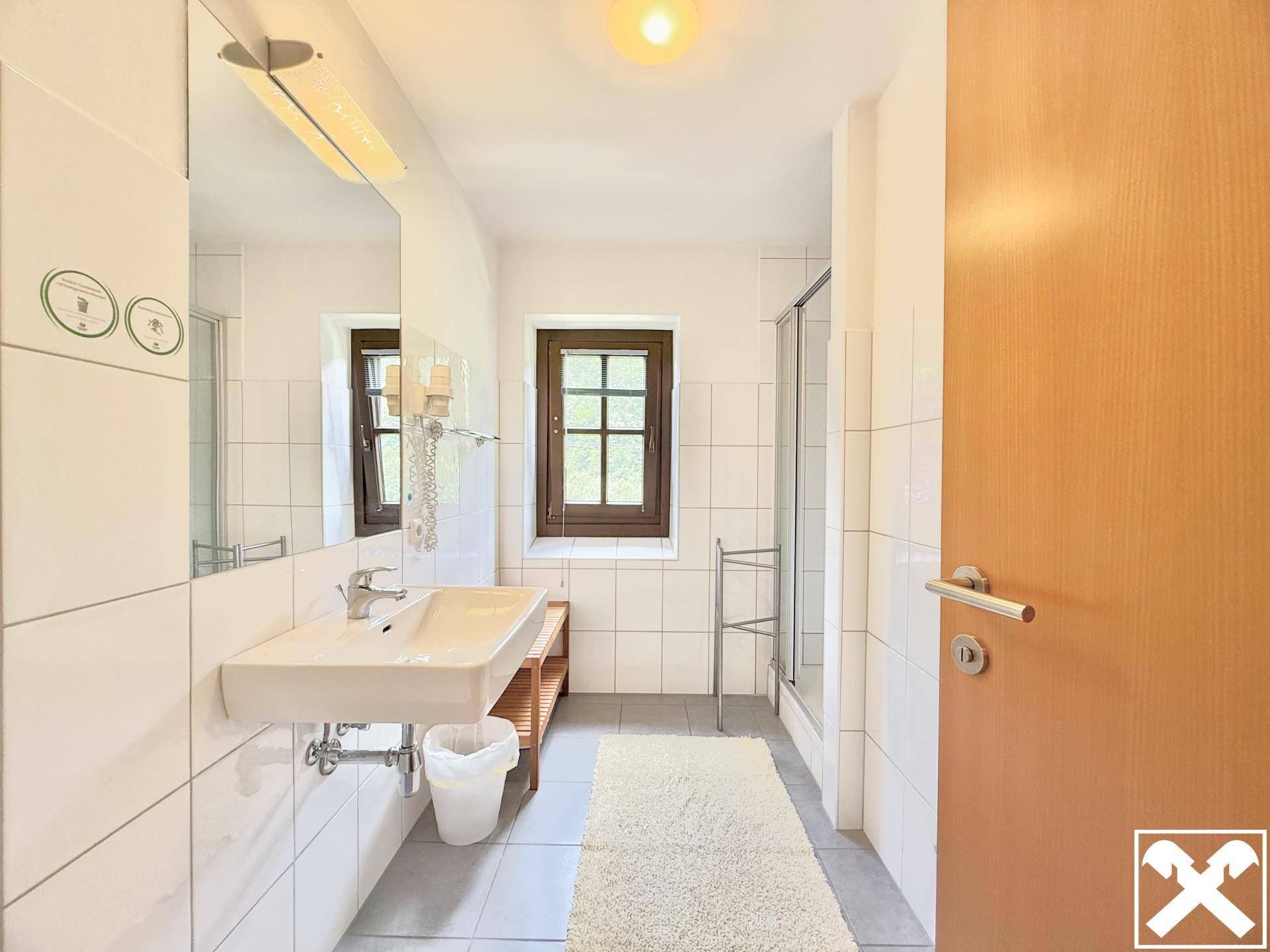 Helles Badezimmer mit weißen Fliesen, Einzelwaschbecken und Duschkabine sowie Fenster.