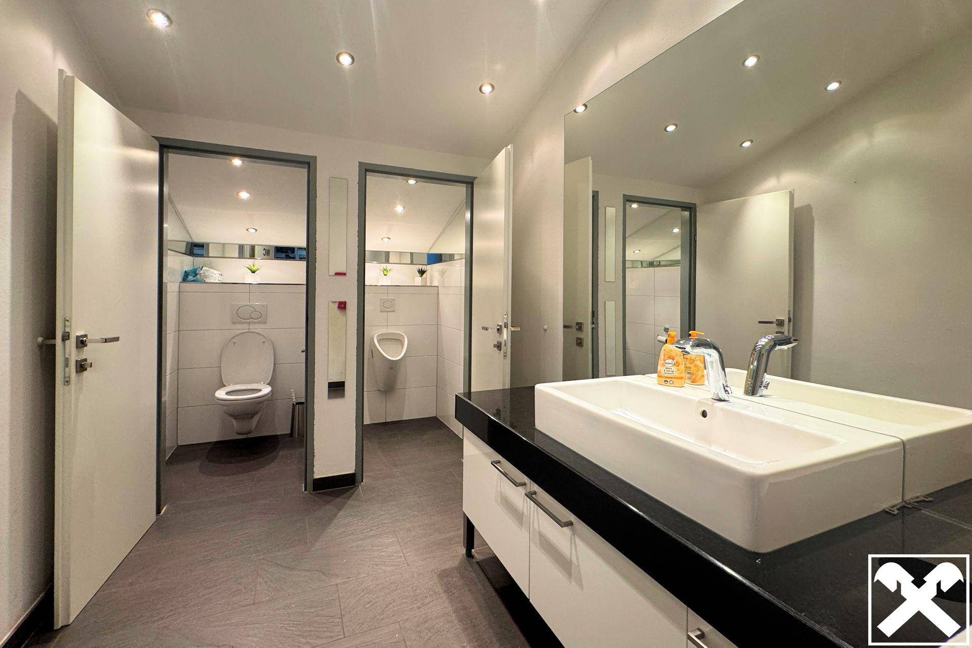 Moderner Sanitärbereich mit mehreren Toilettenkabinen, Urinal und großem Waschbecken mit schwarzer Arbeitsplatte.