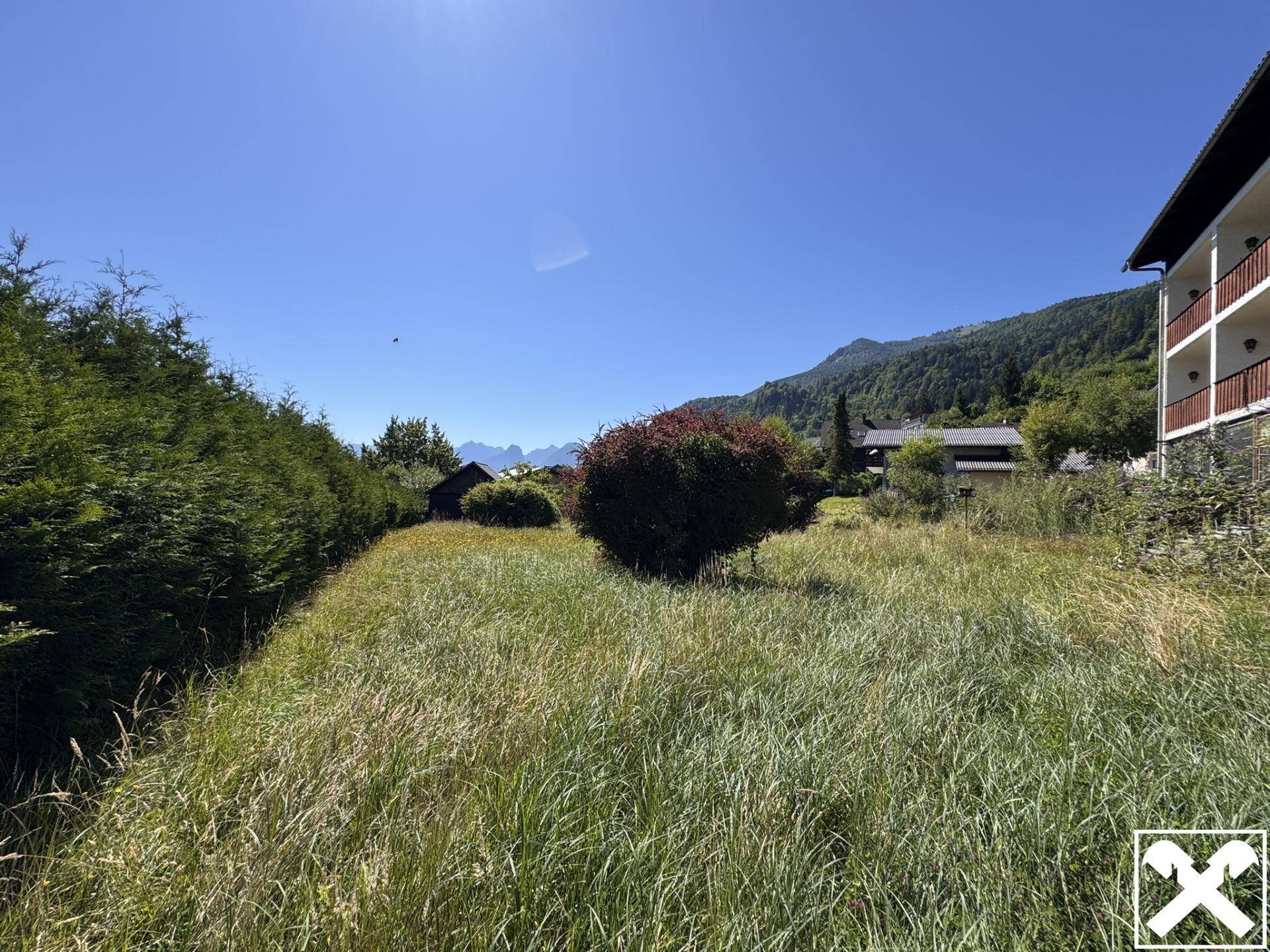 Weitläufige Grünfläche mit Bäumen und Blick auf die umliegende Berglandschaft unter blauem Himmel.