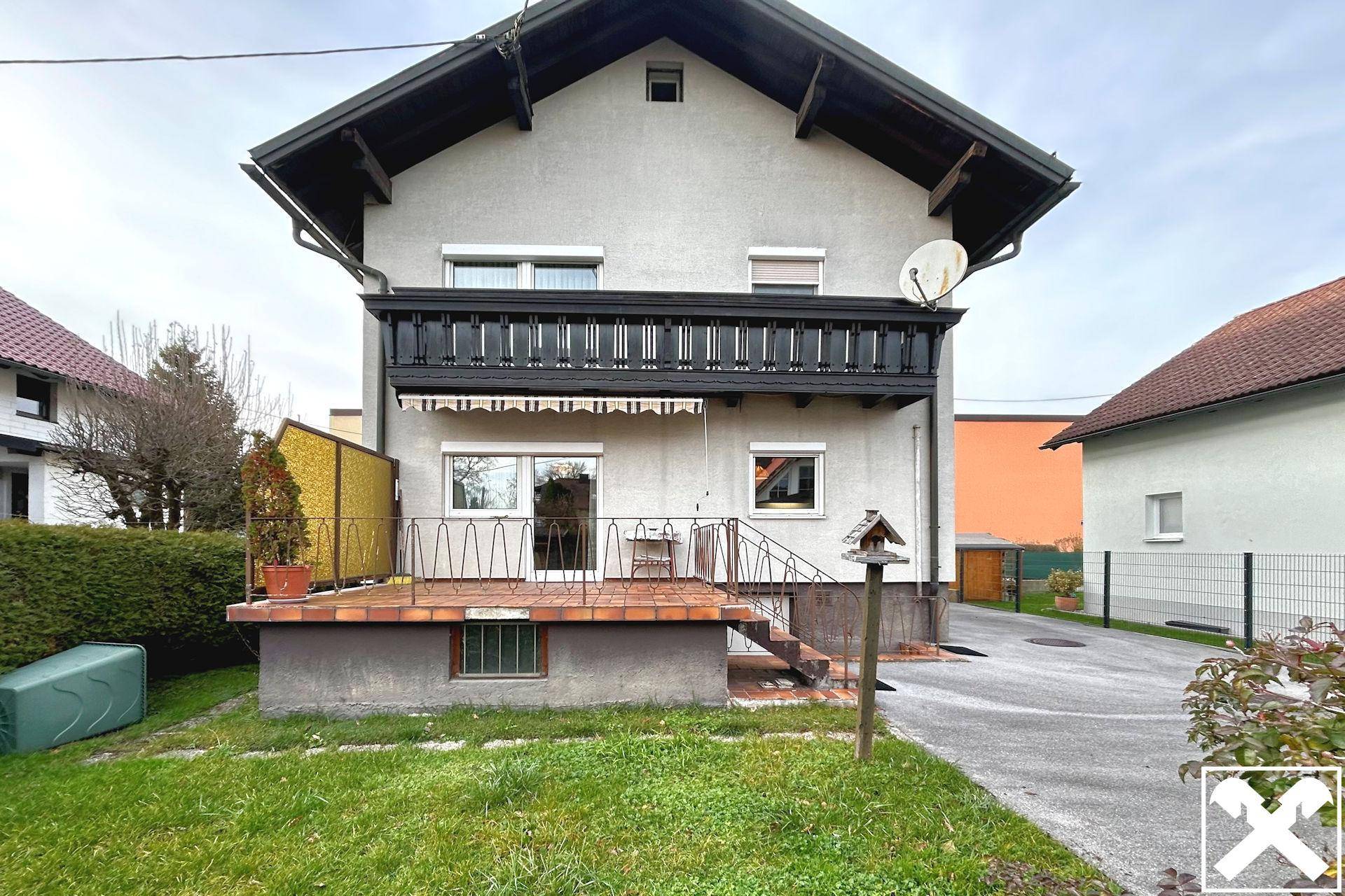 Hausfassade mit Balkon und einer kleinen Terrasse im Gartenbereich, umgeben von Grün.