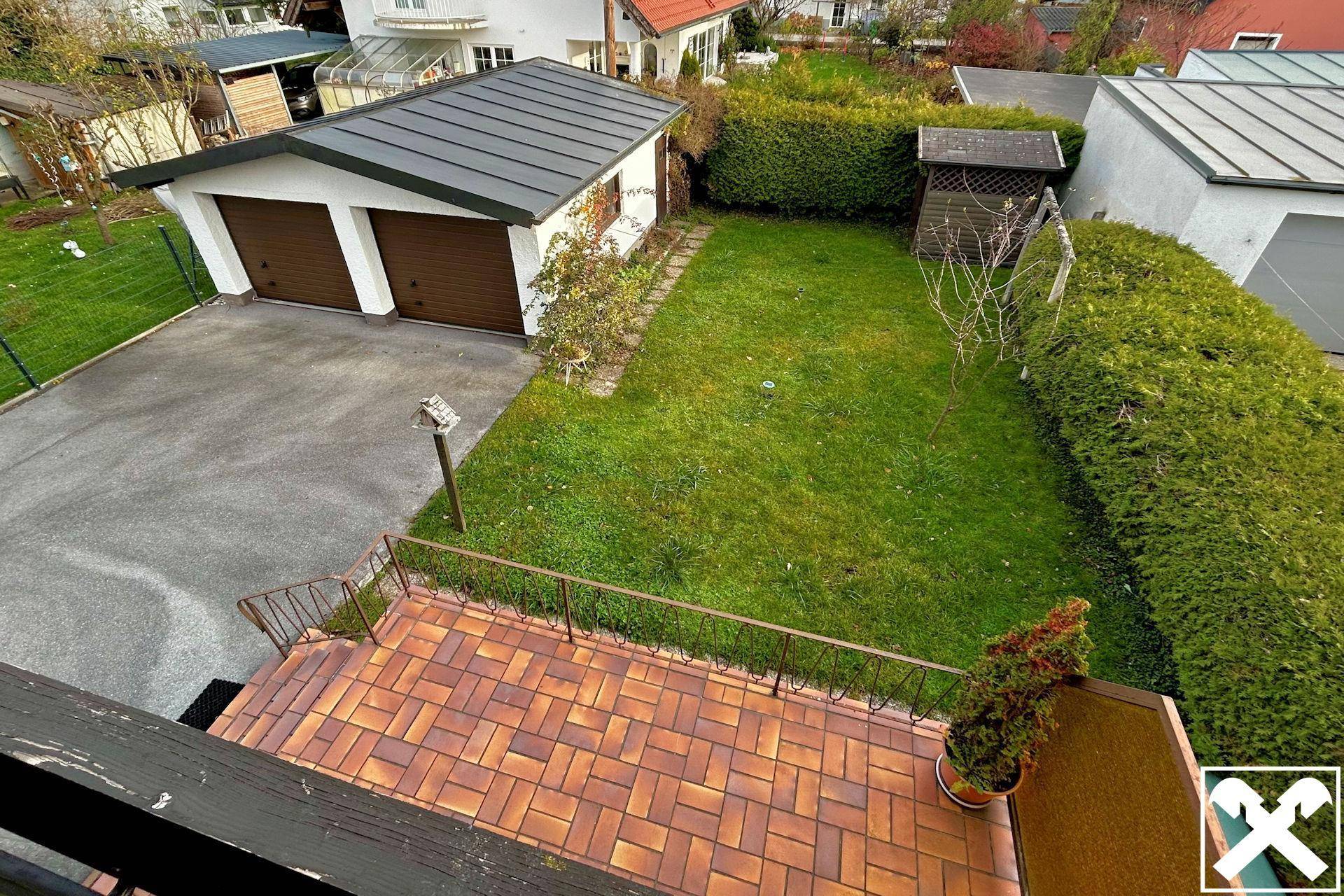 Blick von der Terrasse auf den Garten mit Rasenfläche, Hecken und einer Doppelgarage.
