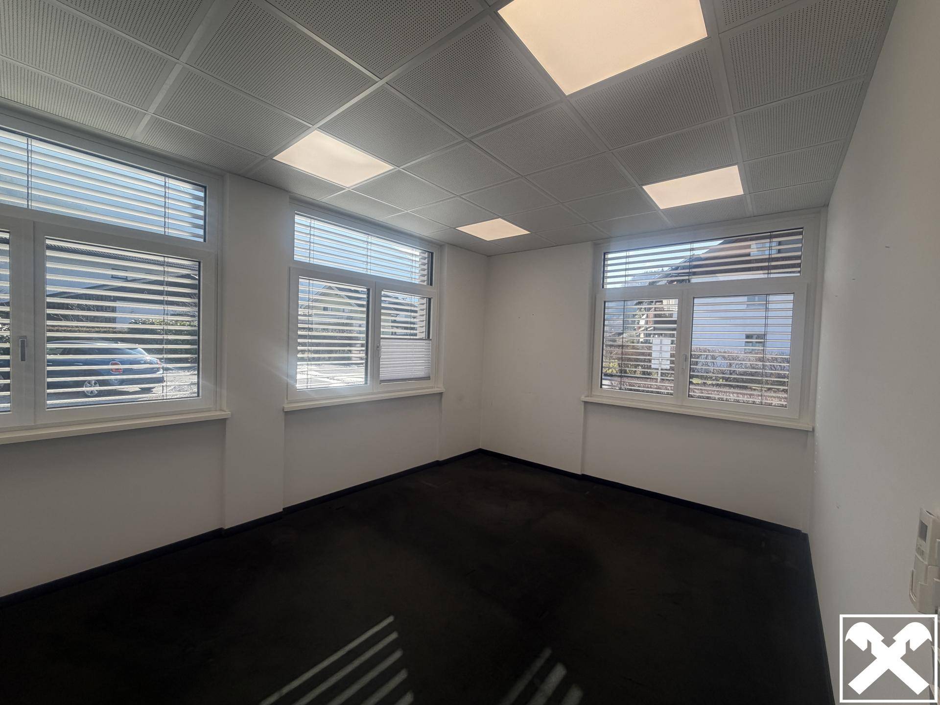 Leeres Büro mit dunklem Boden, mehreren Fenstern mit Jalousien und abgehängter Decke mit quadratischen Leuchten.