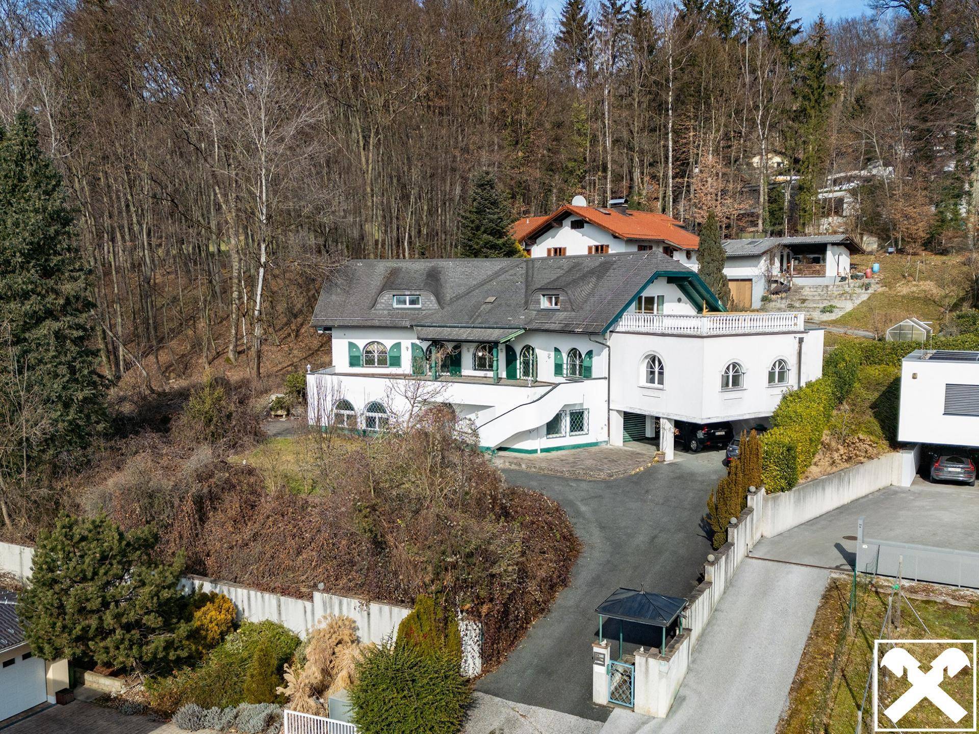 Großzügiges weißes Haus mit grünen Fensterläden und dunklem Dach, eingebettet in eine bewaldete Hanglage.
