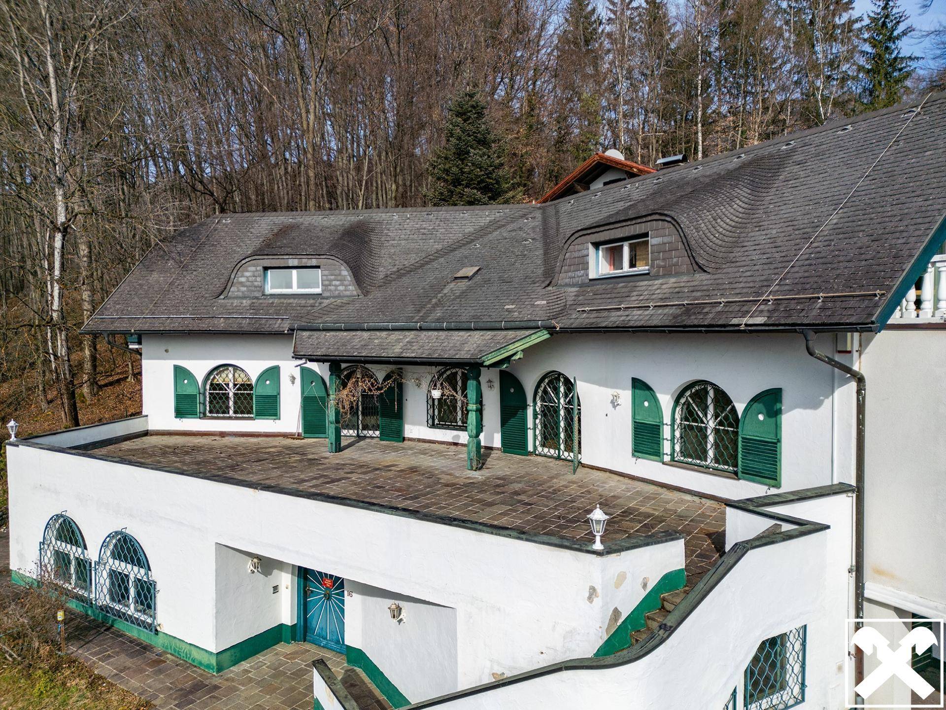 Ansicht des weißen Hauses mit dunklem Dach, grünen Fensterläden und einer weitläufigen Dachterrasse.