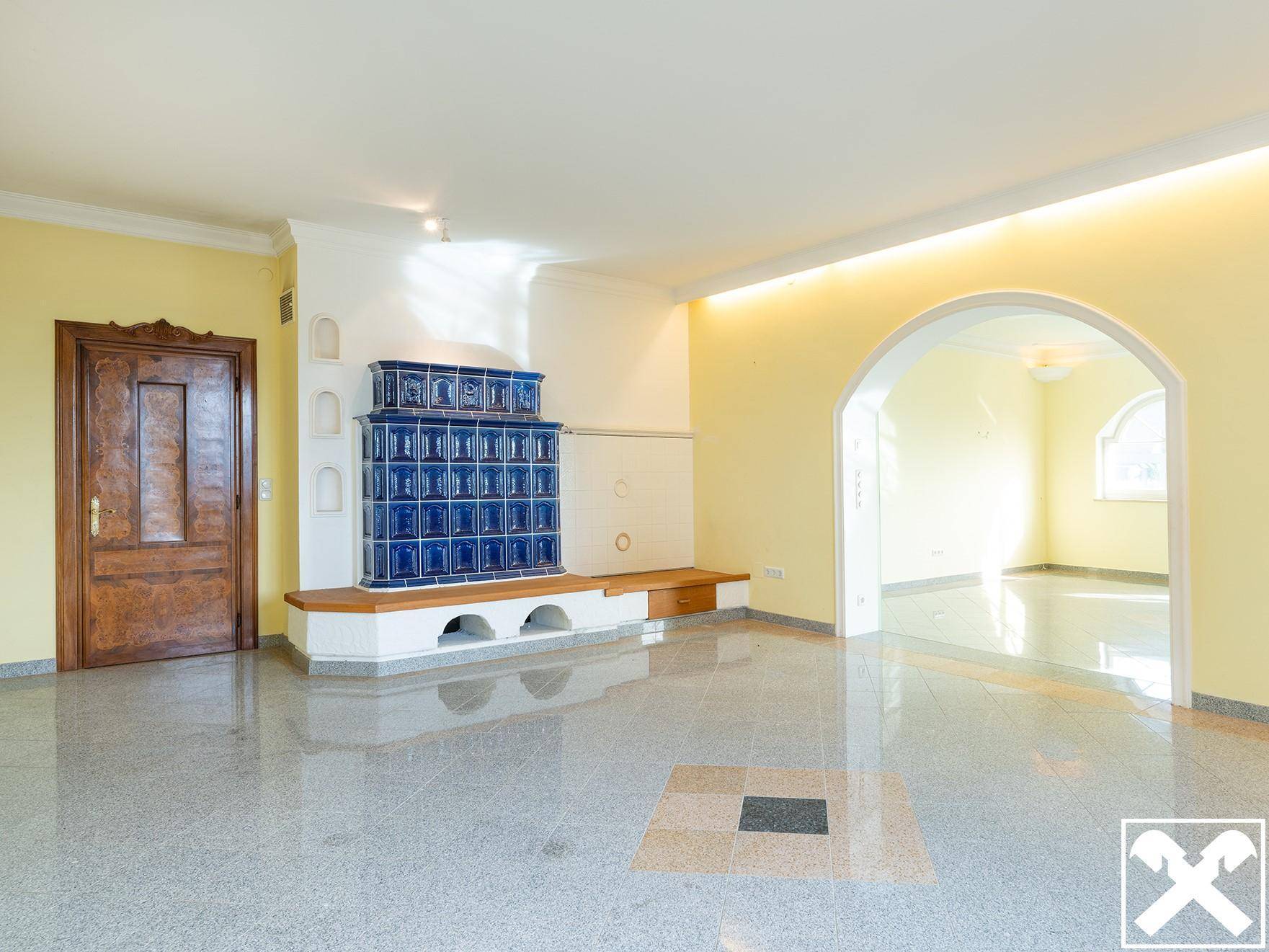 Helles Wohnzimmer mit gelben Wänden, glänzendem Fliesenboden und einem traditionellen blauen Kachelofen.
