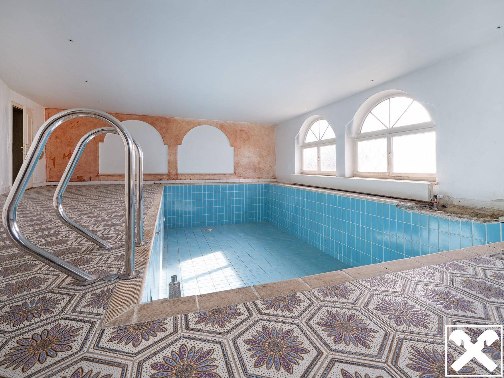 Innenpool mit blauen Fliesen, dekorativem Mosaikboden und großen Bogenfenstern, in Renovierungszustand.