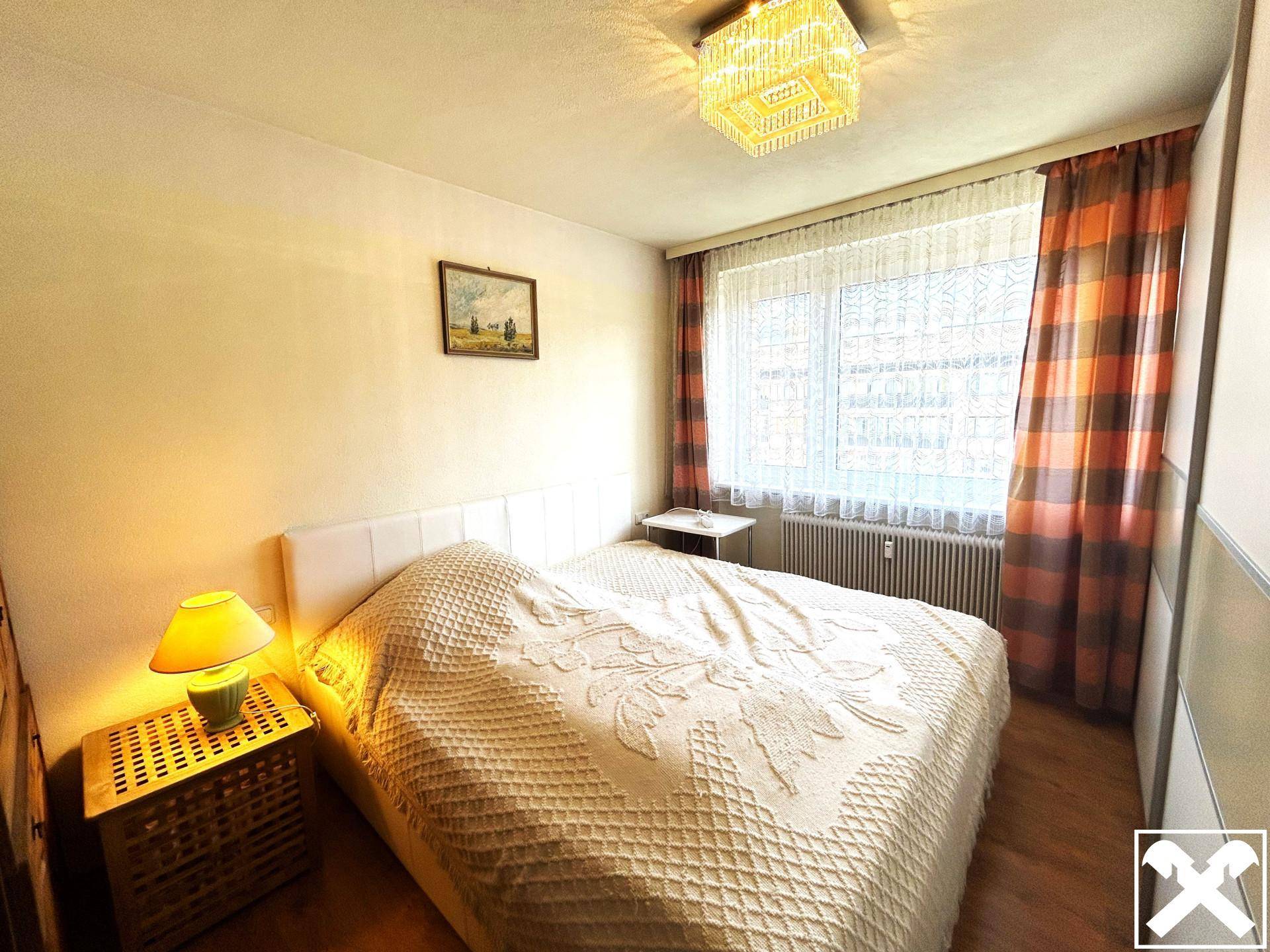 Schlafzimmer mit Doppelbett, Nachttischlampe, Fenster mit Vorhängen und Kleiderschrank.