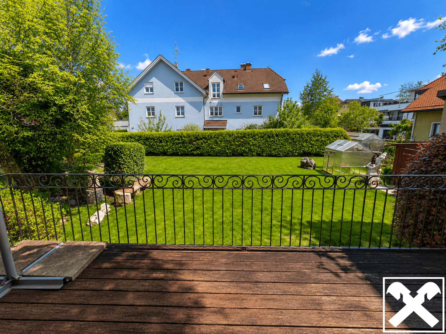 Blick von einem Holzbalkon auf einen weitläufigen, gepflegten Garten mit Hecke und Gewächshaus.