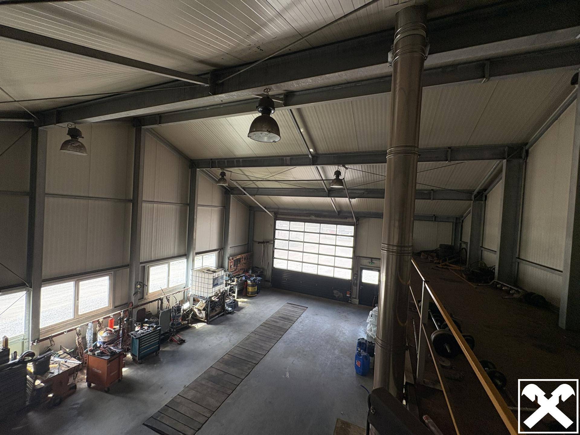 Innenansicht der Industriehalle von oben, mit Werkzeugen, hohen Decken und dem großen Rolltor, das Tageslicht hereinlässt.