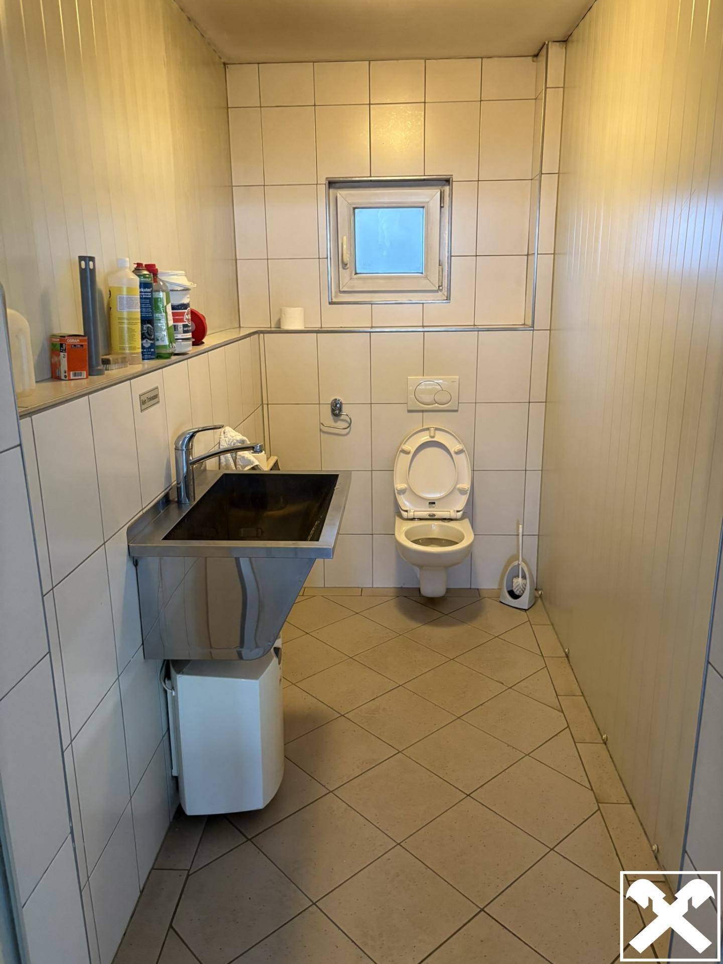 Einfache Toilette mit weißen Fliesen, einem WC und einem Waschbecken aus Edelstahl, ausgestattet mit einem Fenster.