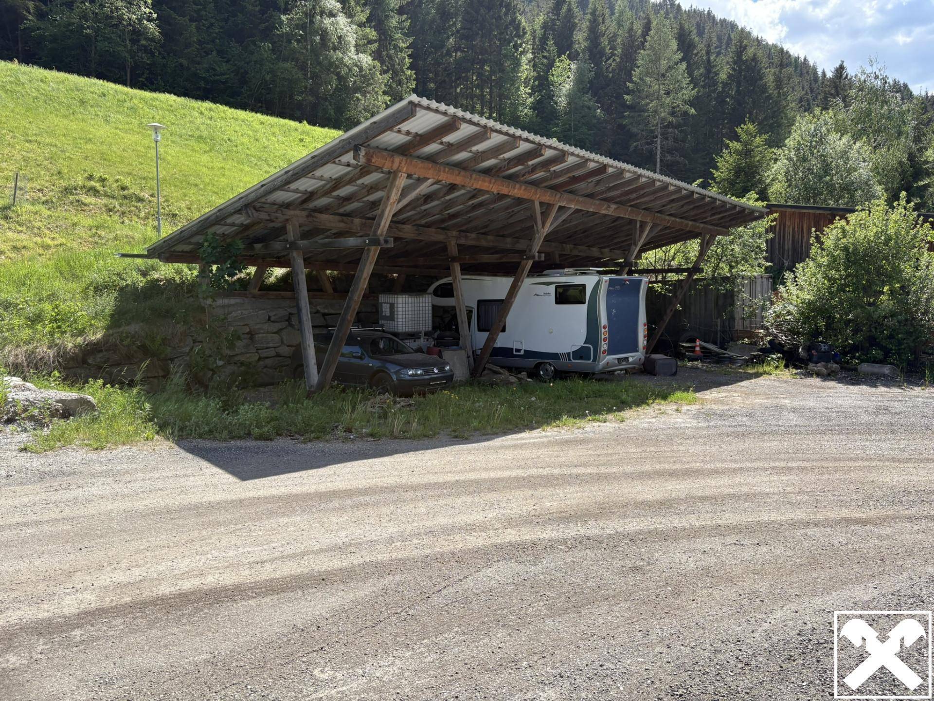 Rustikaler Holz-Carport bietet Schutz für ein Auto und einen Wohnwagen, eingebettet in eine grüne Hügellandschaft.
