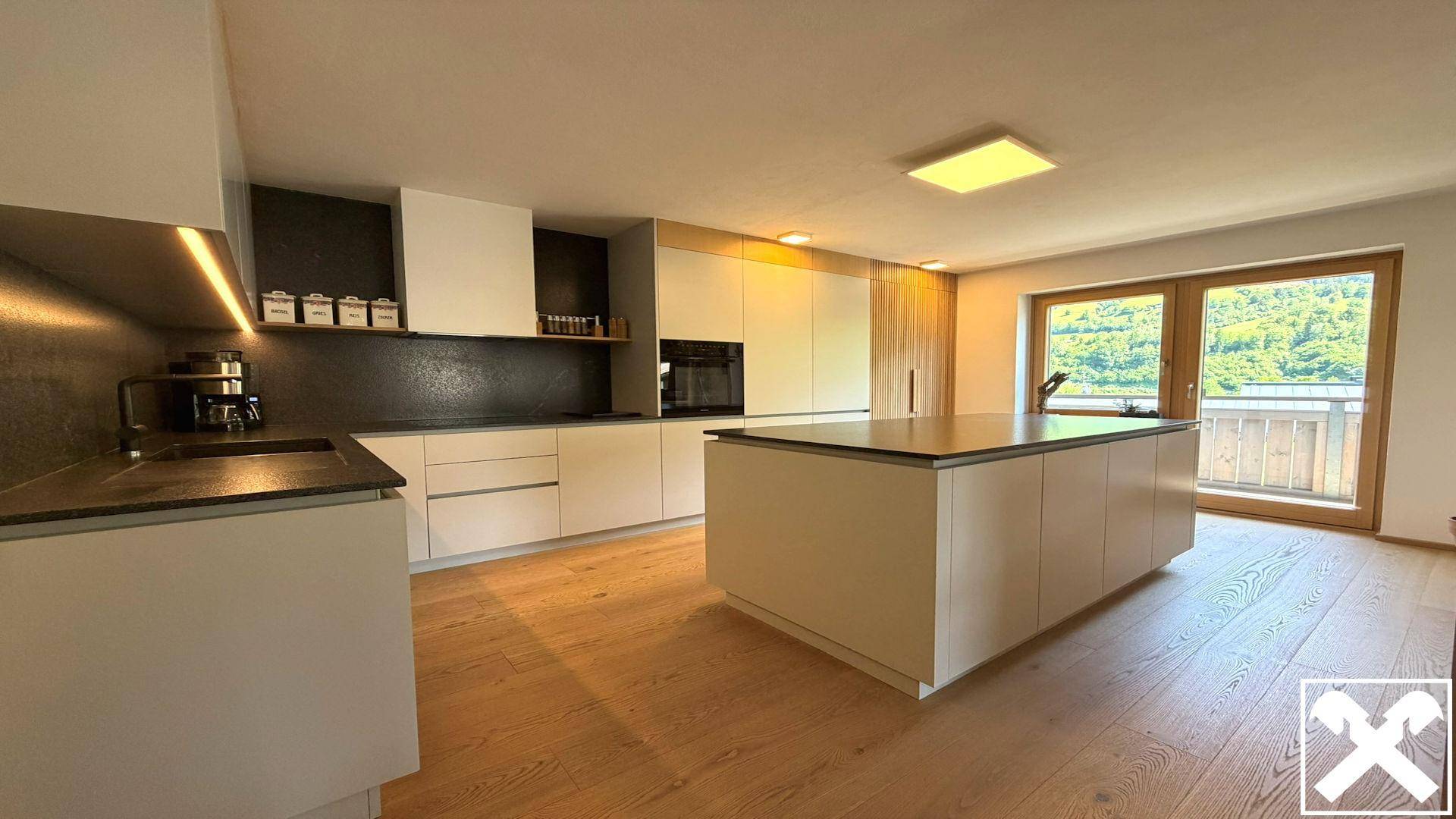 Moderne Küche mit Kücheninsel, hellen Fronten und Zugang zu einem Balkon mit Bergblick.