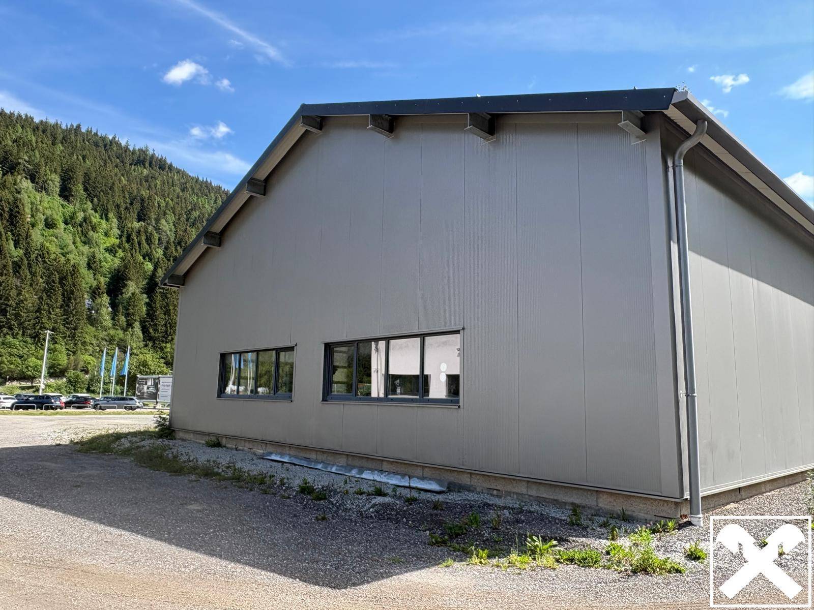 Seitenansicht der Gewerbehalle mit Fenstern und Schotterfläche, umgeben von bewaldeten Bergen unter blauem Himmel.