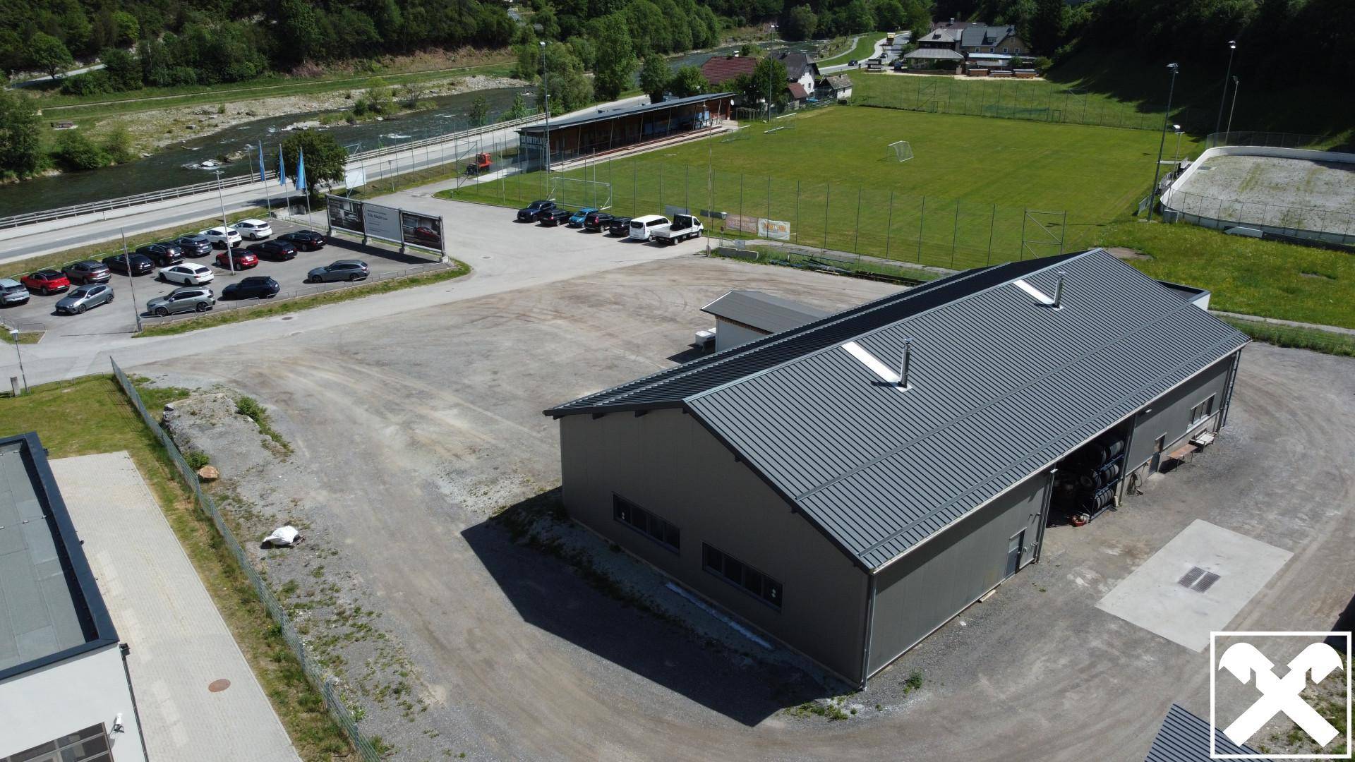 Luftaufnahme der Gewerbehalle mit umliegenden Parkplätzen, einem Fluss und einem Sportplatz im Hintergrund.