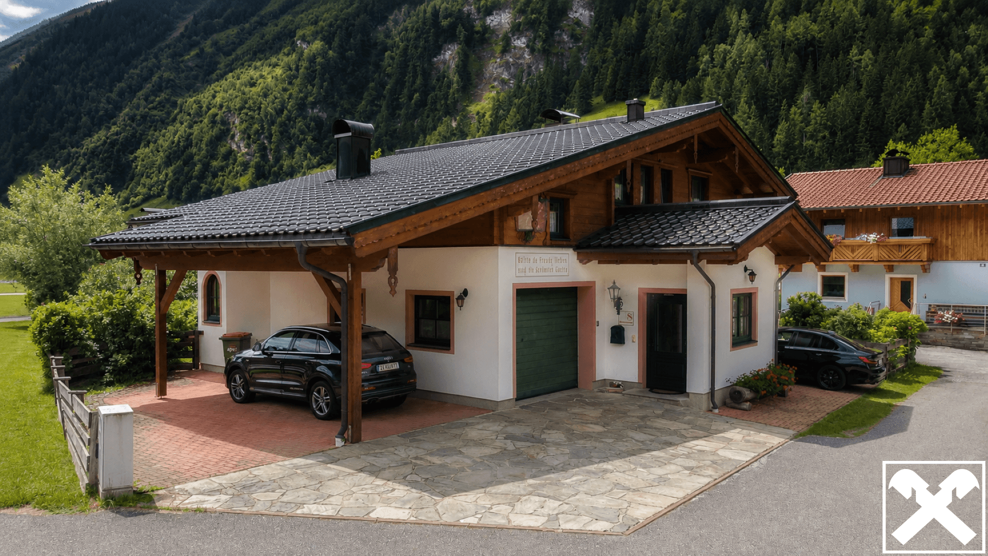 Traditionelles Alpenhaus mit Carport, Garage und gepflasterter Auffahrt vor bewaldeten Bergen.
