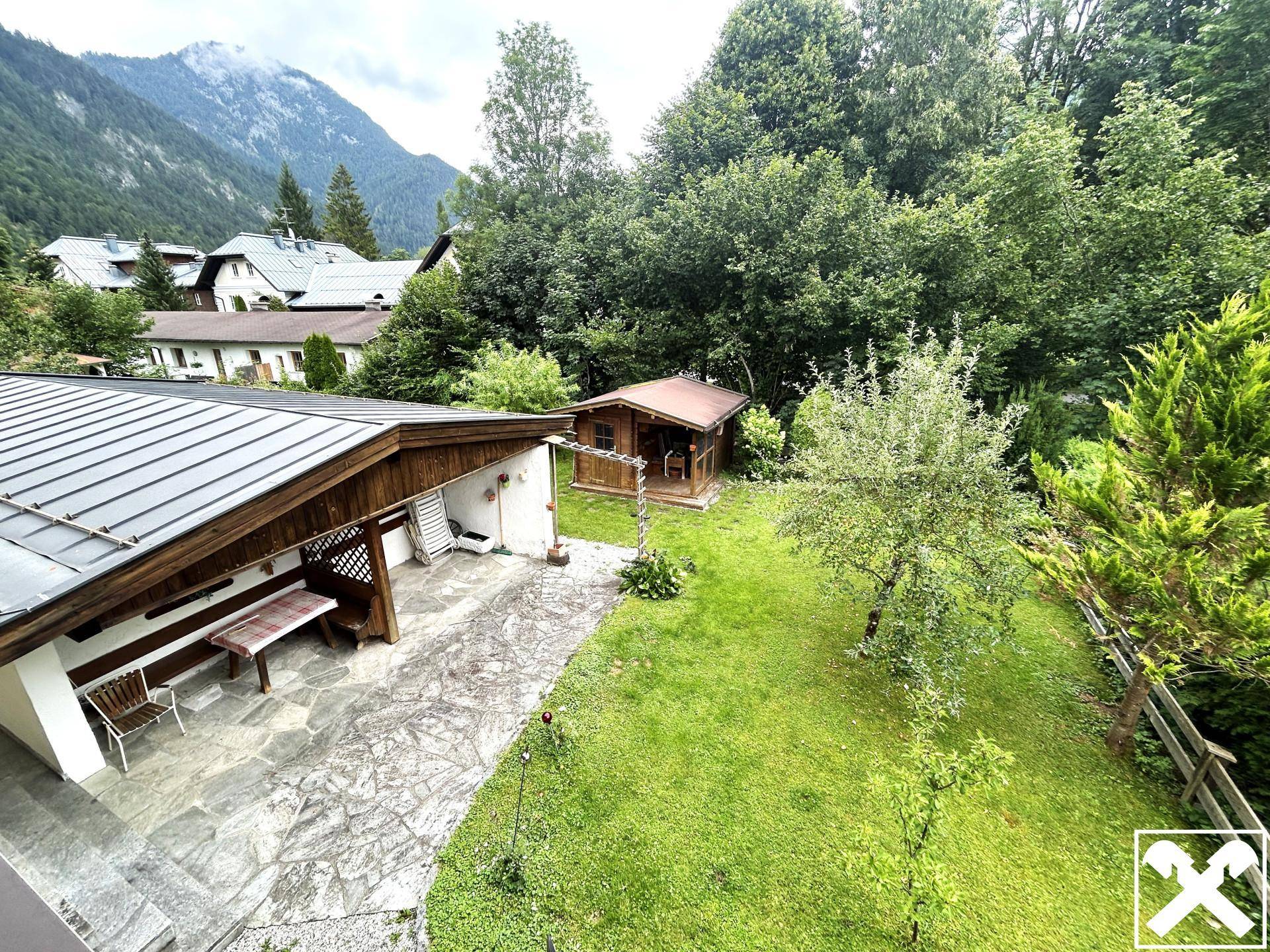 Gartenbereich mit Rasenfläche, gepflastertem Sitzplatz und kleinem Gartenhaus.