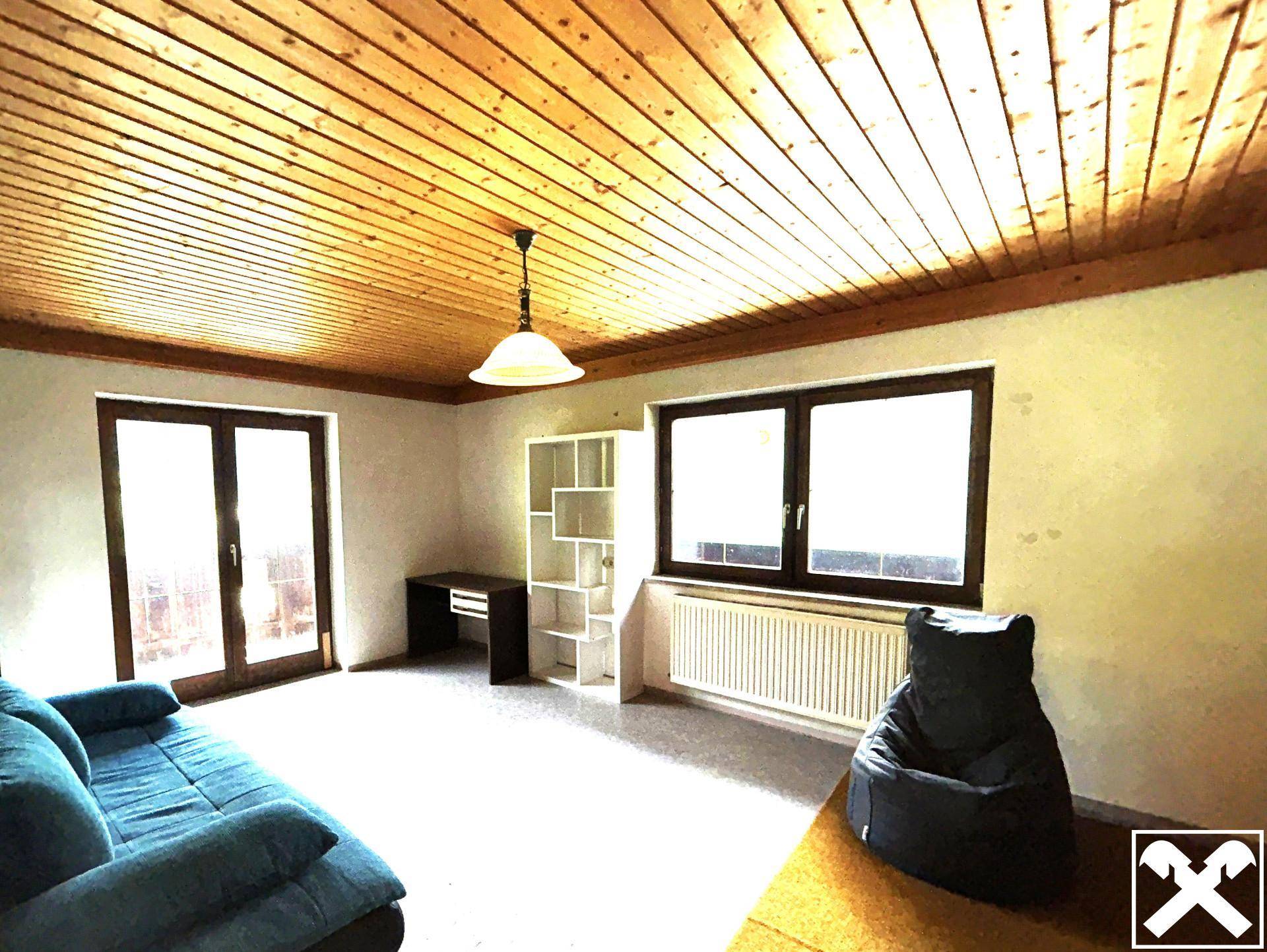 Helles Wohnzimmer mit Holzbalkendecke, großem Fenster und Zugang zu einem Außenbereich.